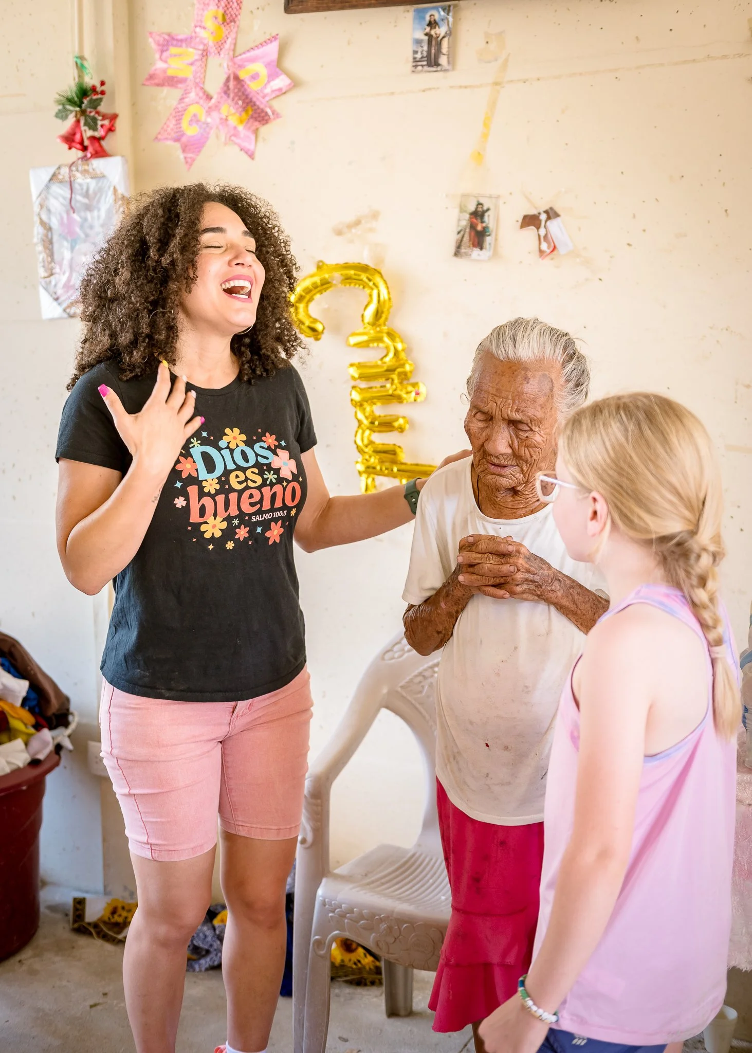 A group of women, including a young girl, are gathered in a room decorated with pink and yellow patriotic banners and a gold balloon. They are joyful and engaged in conversation, with one woman wearing a black t-shirt that says 'Dios es bueno' and ha