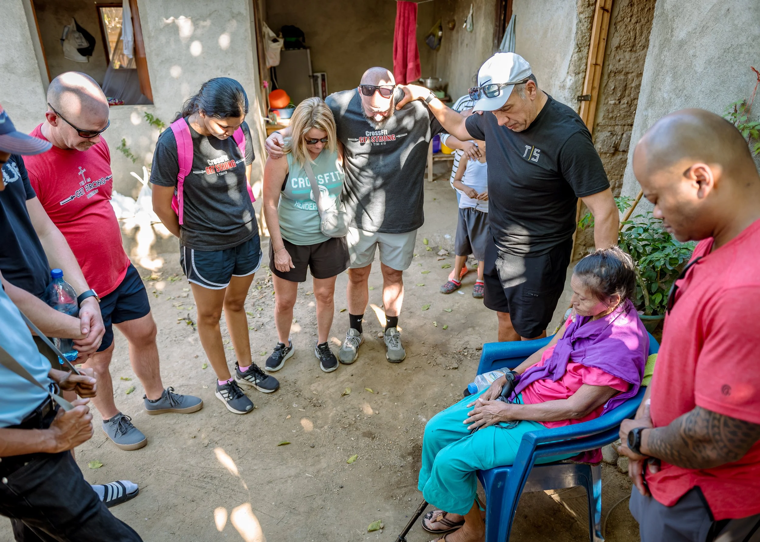 A group of people gather outdoors in a circle, with one woman seated in a blue chair and the rest standing around her with heads bowed, holding hands or placing hands on each other's shoulders in a circle of support and prayer.