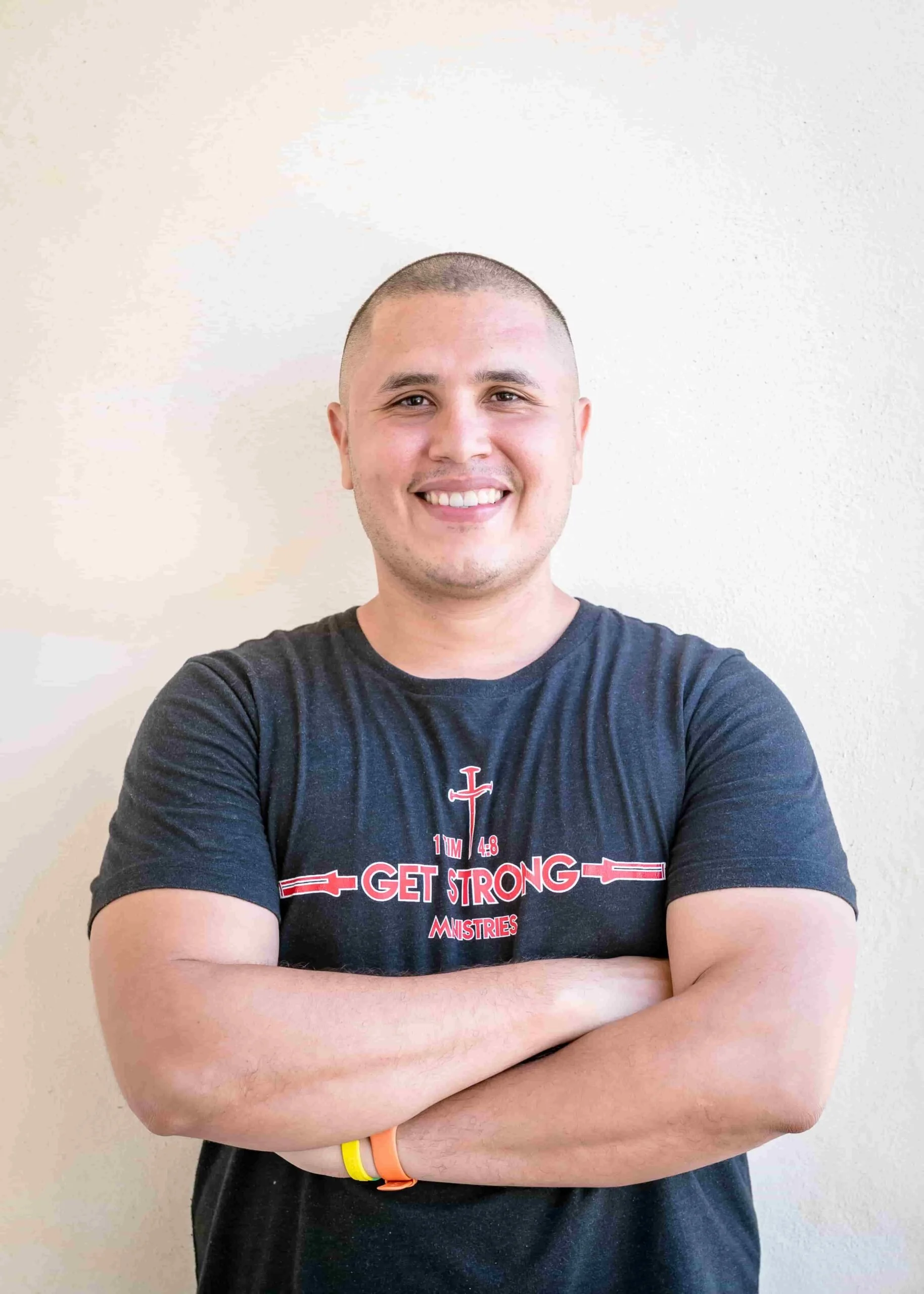 A young man with short hair, smiling, wearing a dark T-shirt with a logo that says "Get Strong Ministries," standing with his arms crossed against a plain, light-colored wall.