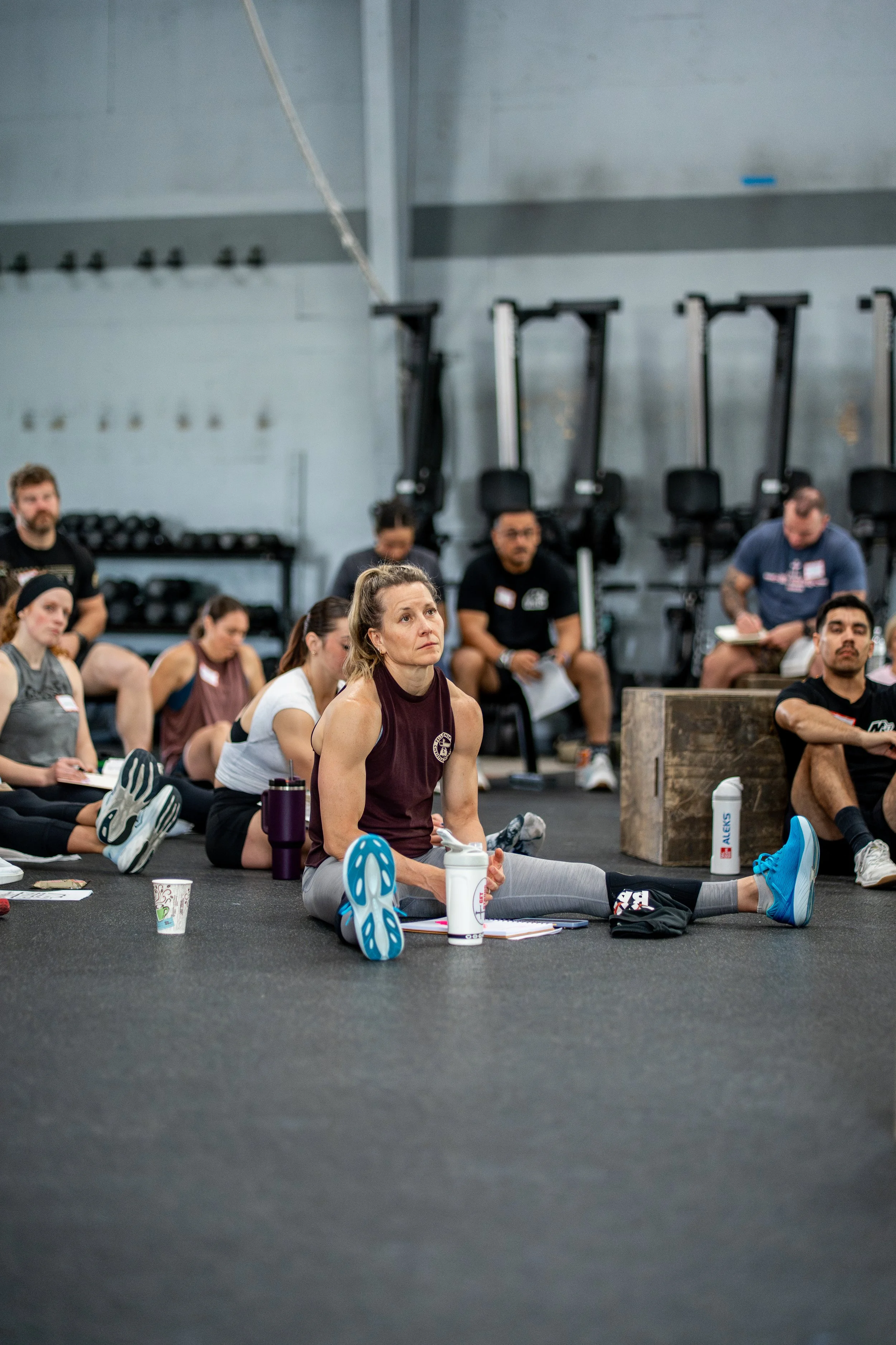 People participating in a fitness class or workshop in a gym or studio, sitting on the floor with fitness equipment around them, listening attentively.