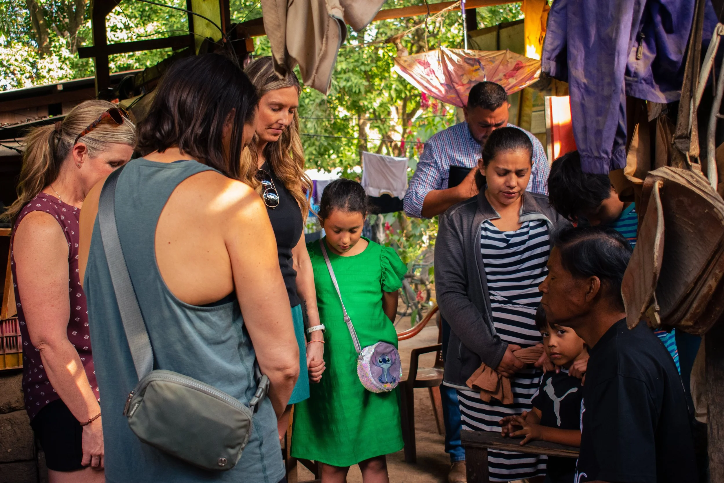 Group of diverse people, including children and adults, gathered around a table outdoors, engaged in a shared activity under a wooden roof with trees and sunlight in the background.
