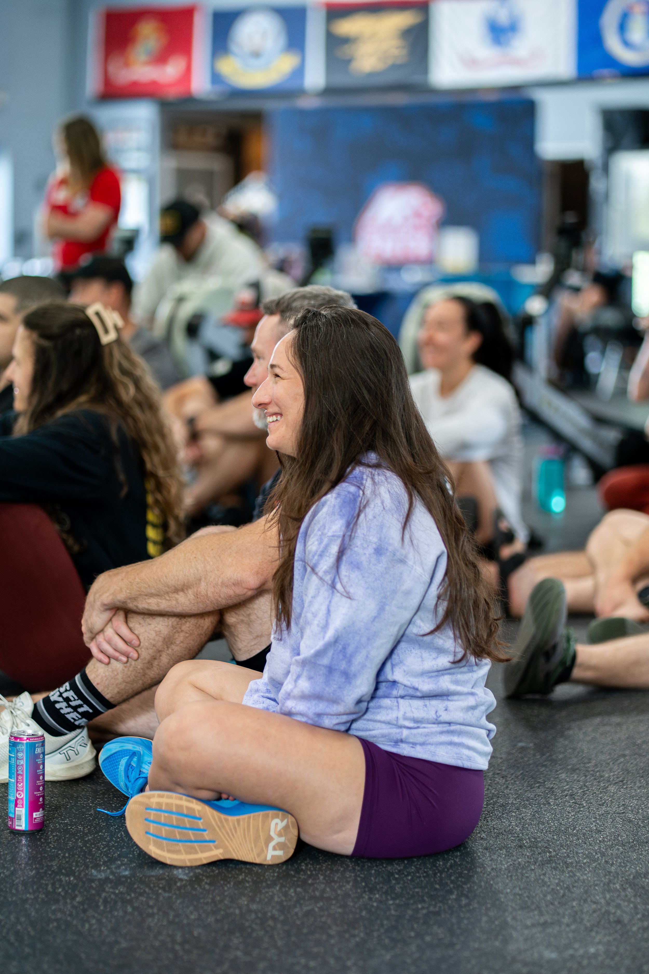 A group of people sitting on the floor during a fitness class in a gym. The focus is on a young woman with long hair, smiling, wearing a purple shirt and shorts, sitting cross-legged.