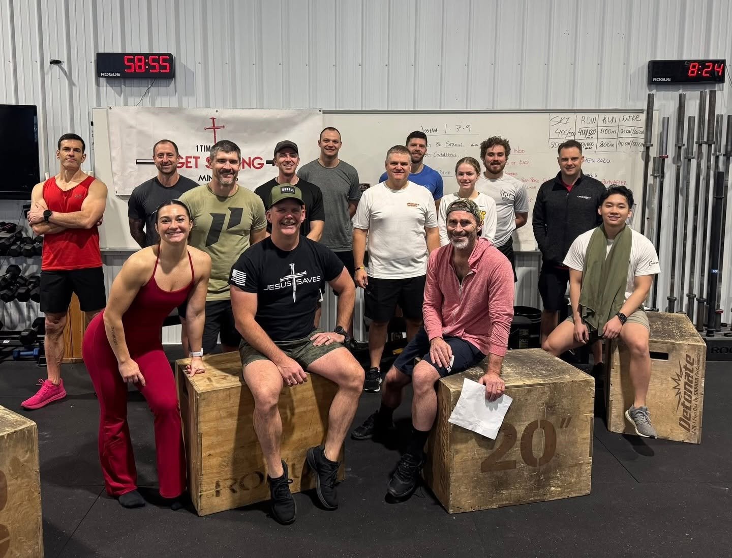 Group of people posing in a gym, some sitting on wooden boxes, others standing behind them, with workout equipment and a whiteboard in the background.