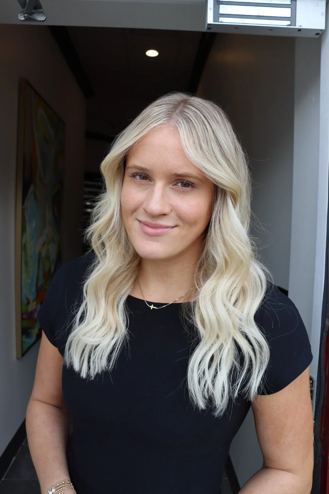 A young woman with long, wavy blonde hair and a black top standing indoors near a doorway.