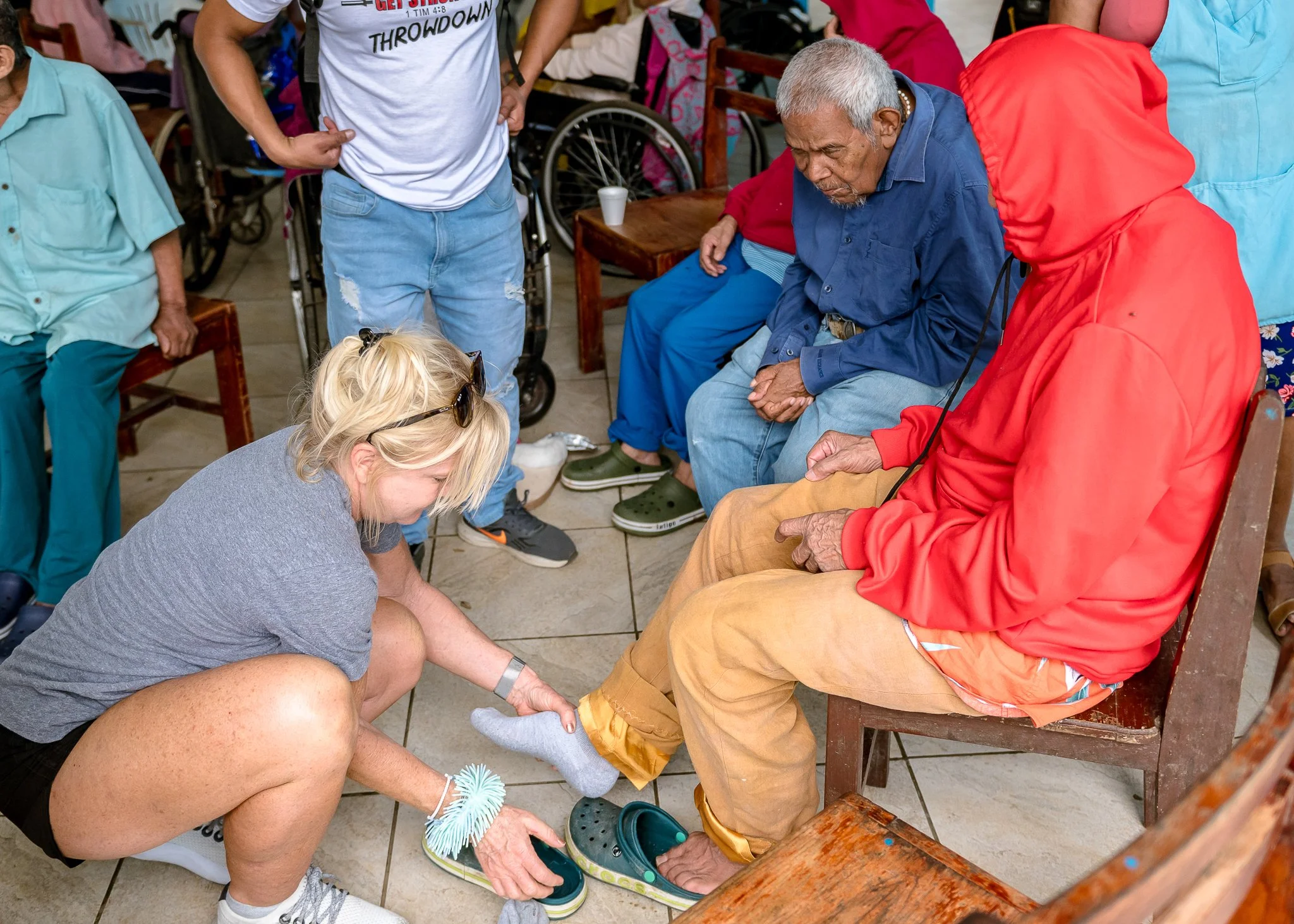 People gathered around, with some sitting and others standing, as a woman is helping an elderly man put on a shoe in a social setting. The scene includes some wheelchairs, chairs, and a table, indicating a communal environment.