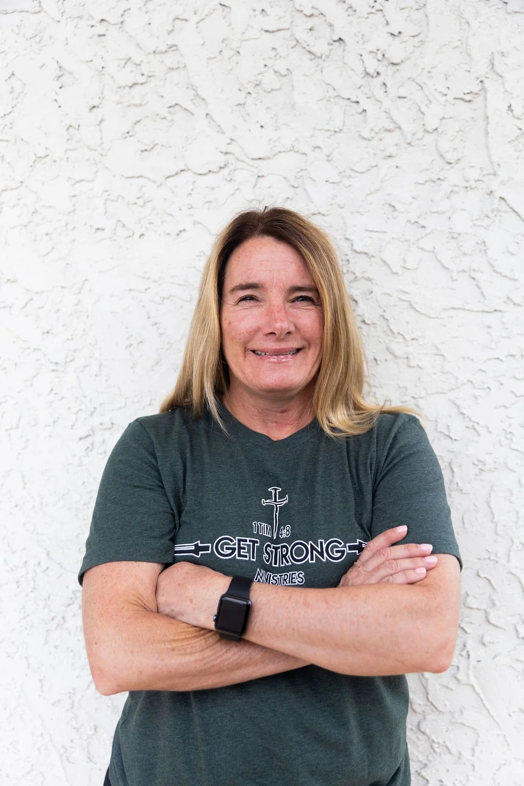 A woman with shoulder-length blonde hair smiling, crossing her arms, wearing a dark green T-shirt with religious text and a cross, standing against a white textured wall.