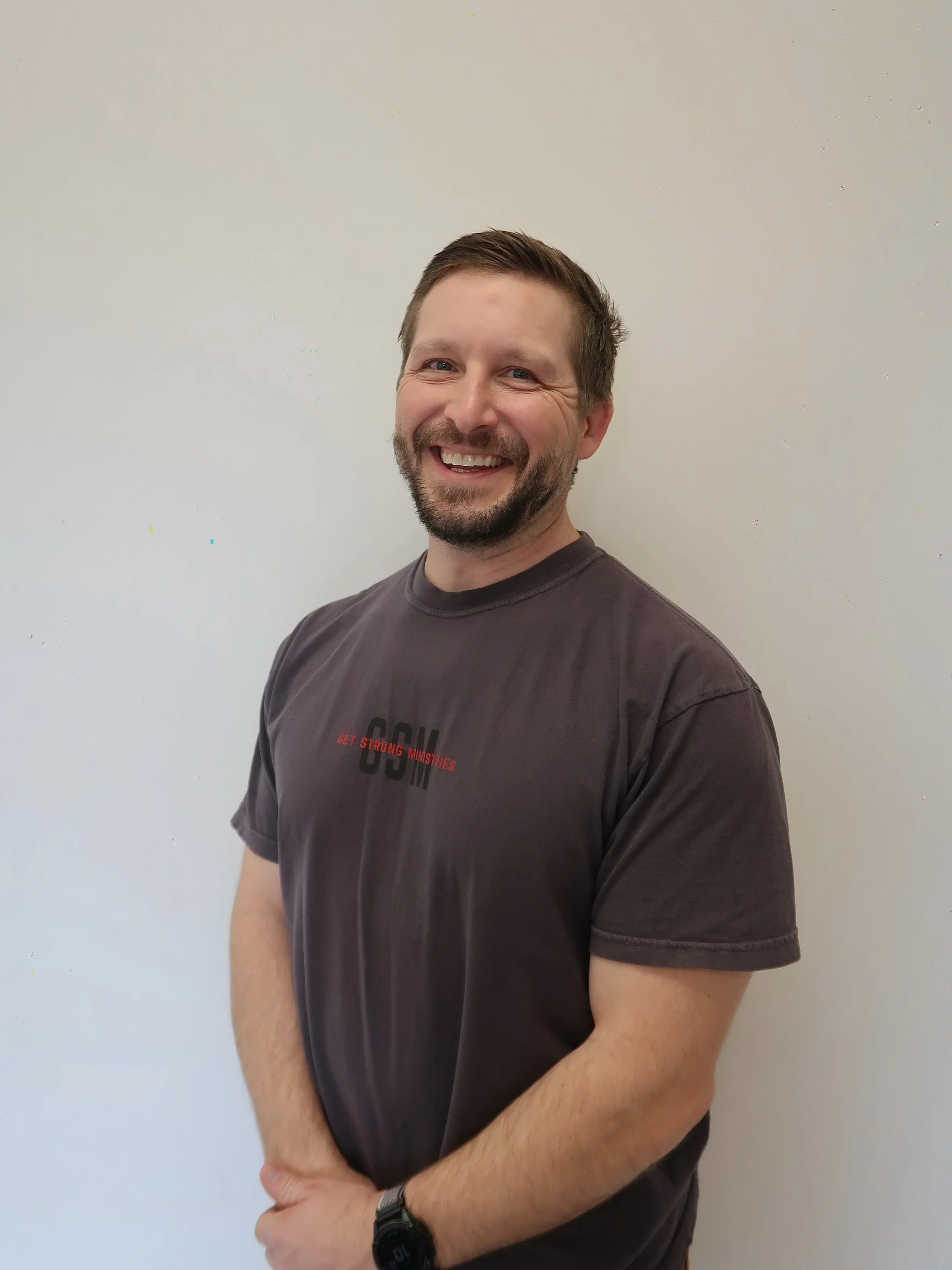 Smiling man with beard wearing a gray t-shirt and black watch standing against a plain light-colored wall.