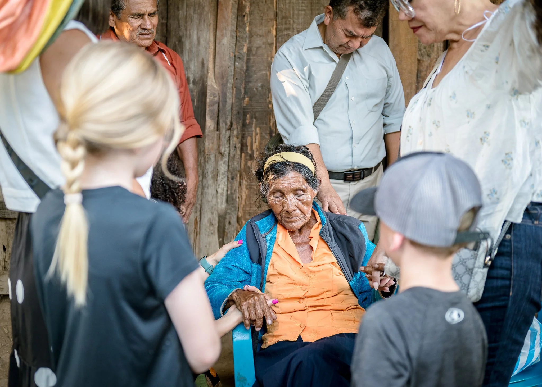A group of people gathered outdoors around an elderly woman, who has her eyes closed, possibly receiving a blessing or prayer to comfort her.