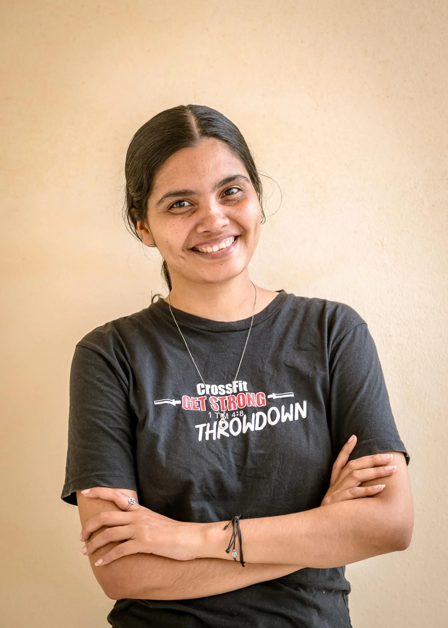 A woman with dark hair smiling and crossing her arms, wearing a black T-shirt with text about CrossFit and a necklace.
