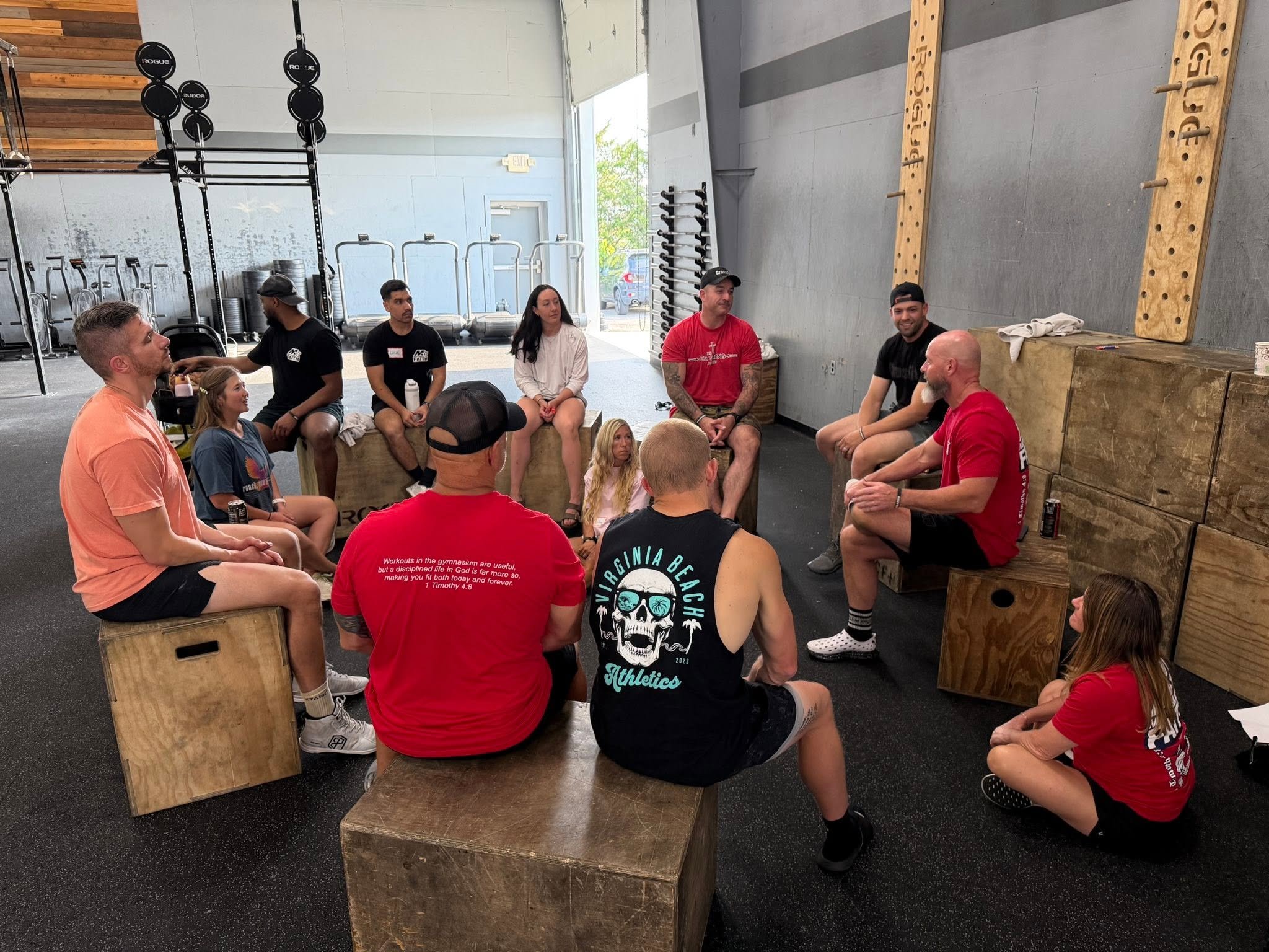 Group of people gathered in a gym or CrossFit gym, sitting on wooden boxes and benches, listening to a man with a beard in a red shirt speaking. The background shows exercise equipment like weights, barbells, and plyometric boxes, with some wooden wall accents and an open garage-style door letting in natural light.