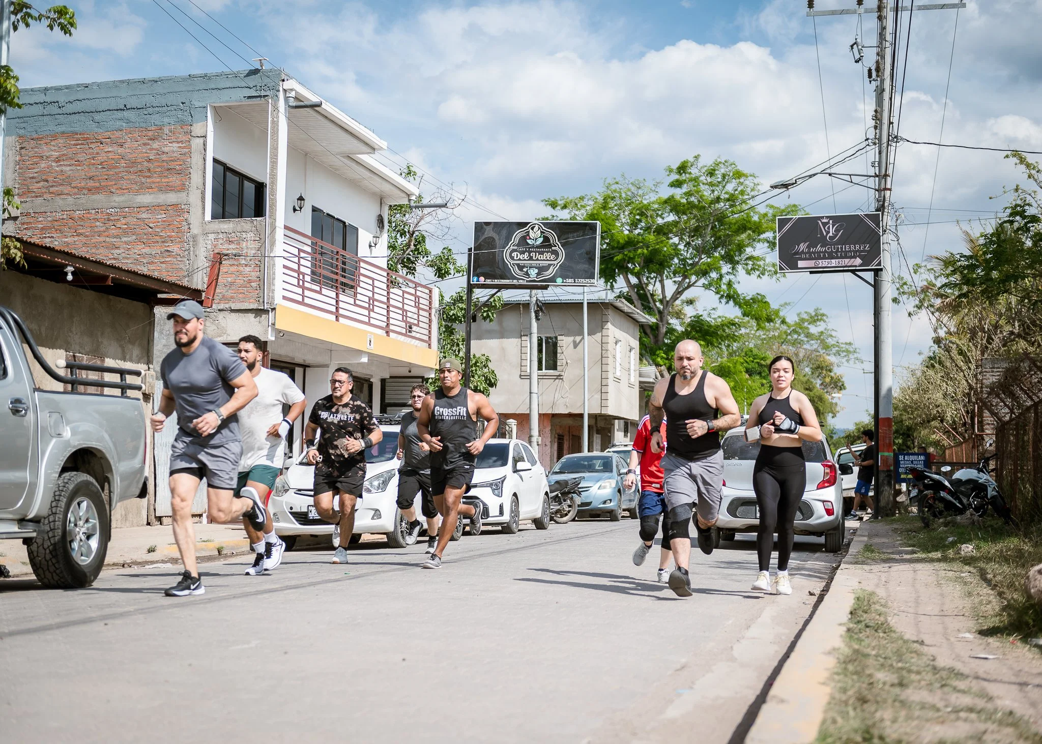 Group of people jogging down a street in a neighborhood with houses, trees, and parked cars. Two billboards are visible, one advertising 'Del Valle' cafe and restaurant, and the other for 'Marlia Gutierrez Beauty Studio.' The sky is partly cloudy.