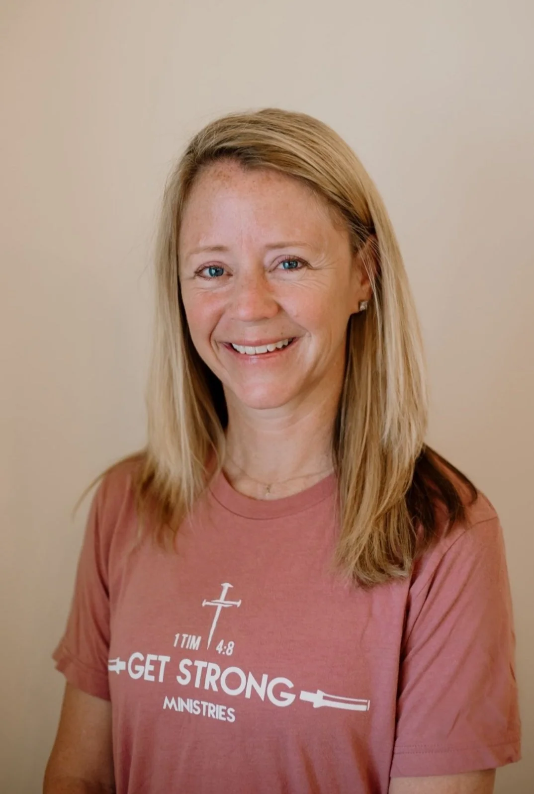A woman with blonde hair and blue eyes smiling, wearing a pink T-shirt that says 'GET STRONG MINISTRIES' with a cross and sword graphic, standing against a plain beige wall.