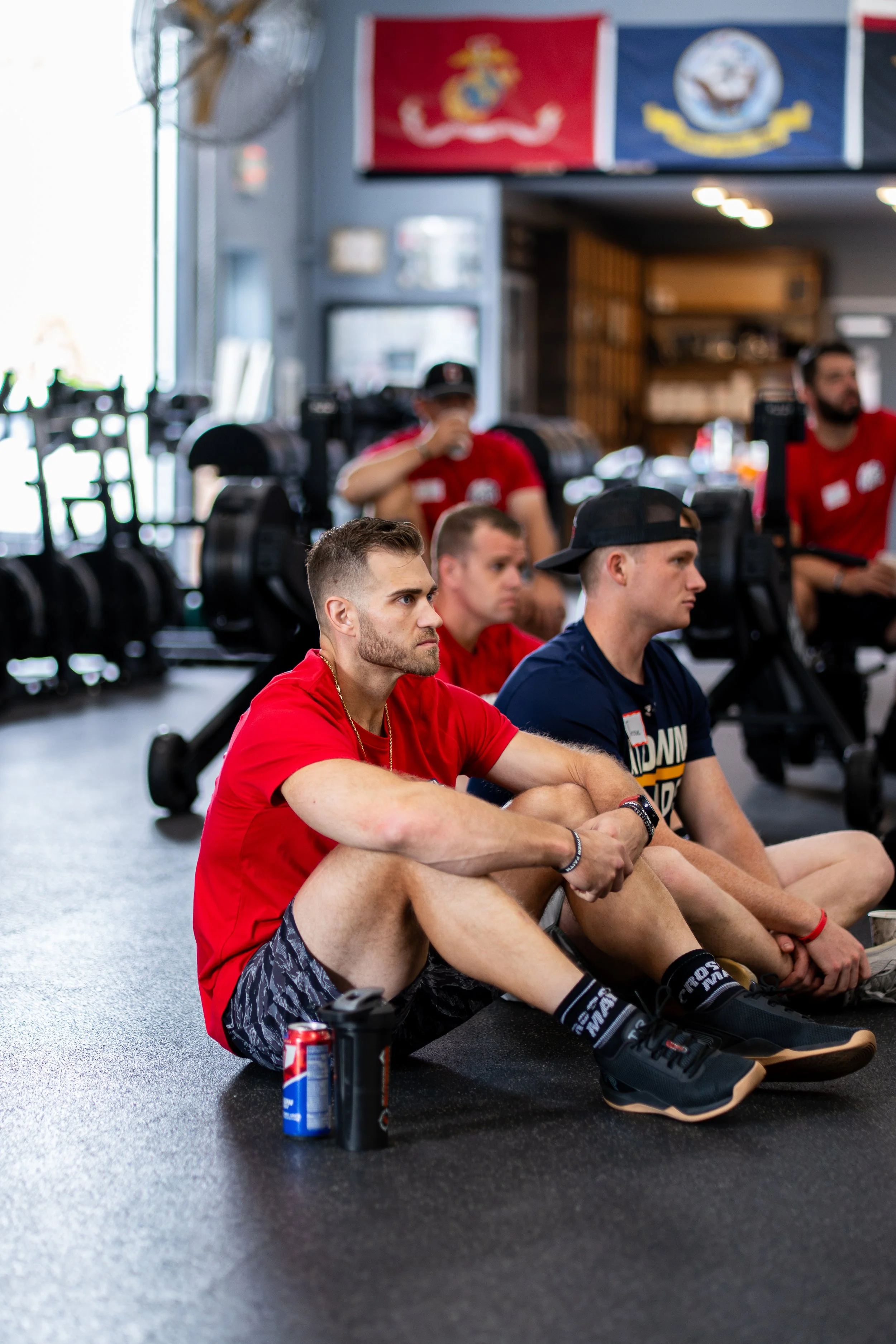 Group of young men sitting on the floor of a gym, listening attentively during a fitness class, with gym equipment in the background.