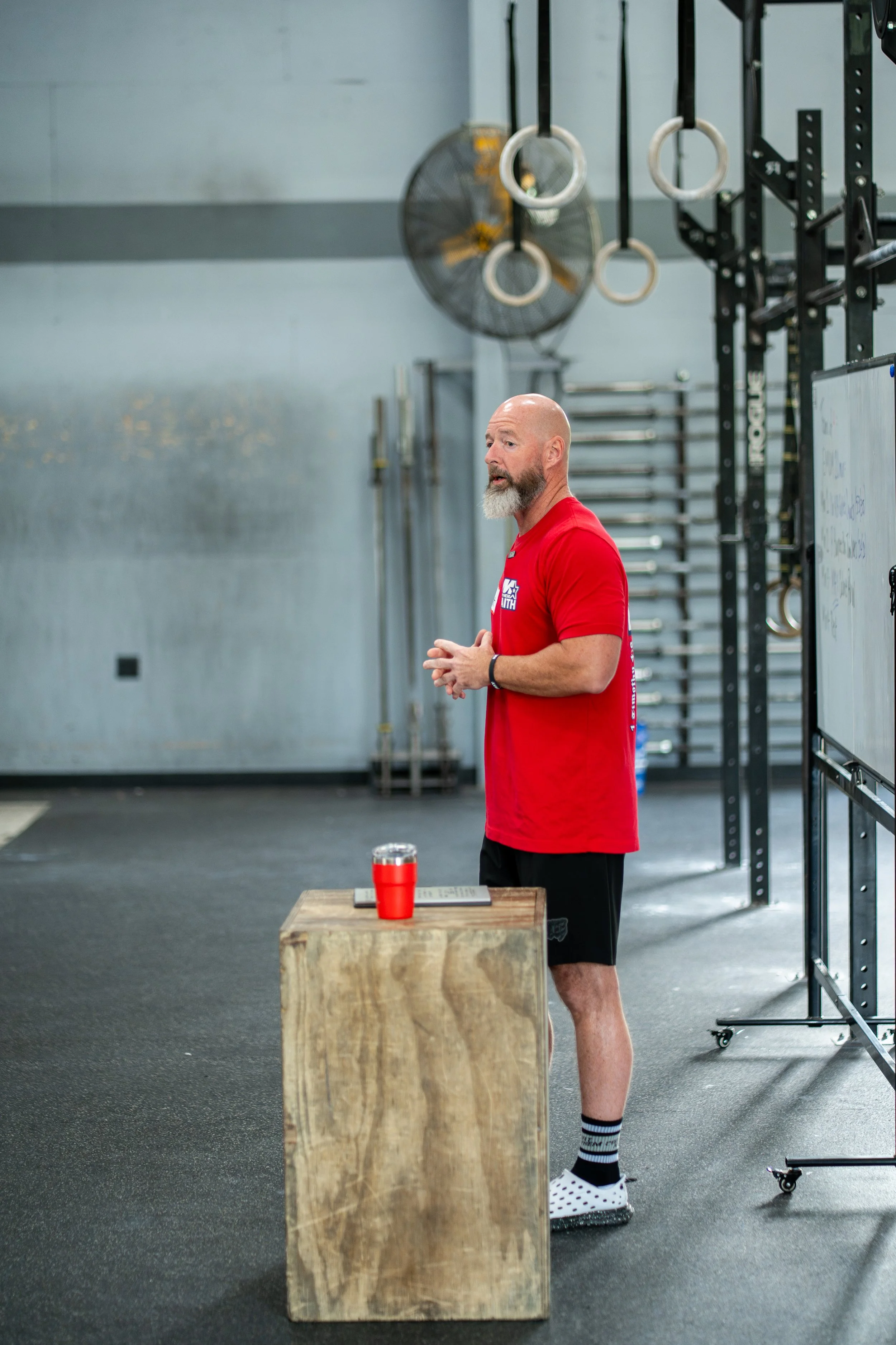 A man with a bald head, beard, and mustache, wearing a red t-shirt, black shorts, and polka dot socks, standing in a gym beside a wooden box, with colorful gymnastic rings hanging from the ceiling and a large industrial fan in the background.