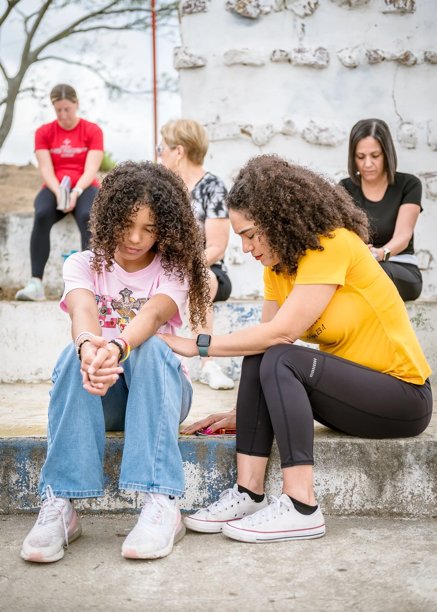 Two young women sit on a step, holding hands and looking down. Two women sit behind them, looking down or away, against a stone wall outdoors. The scene suggests a moment of reflection or comfort.