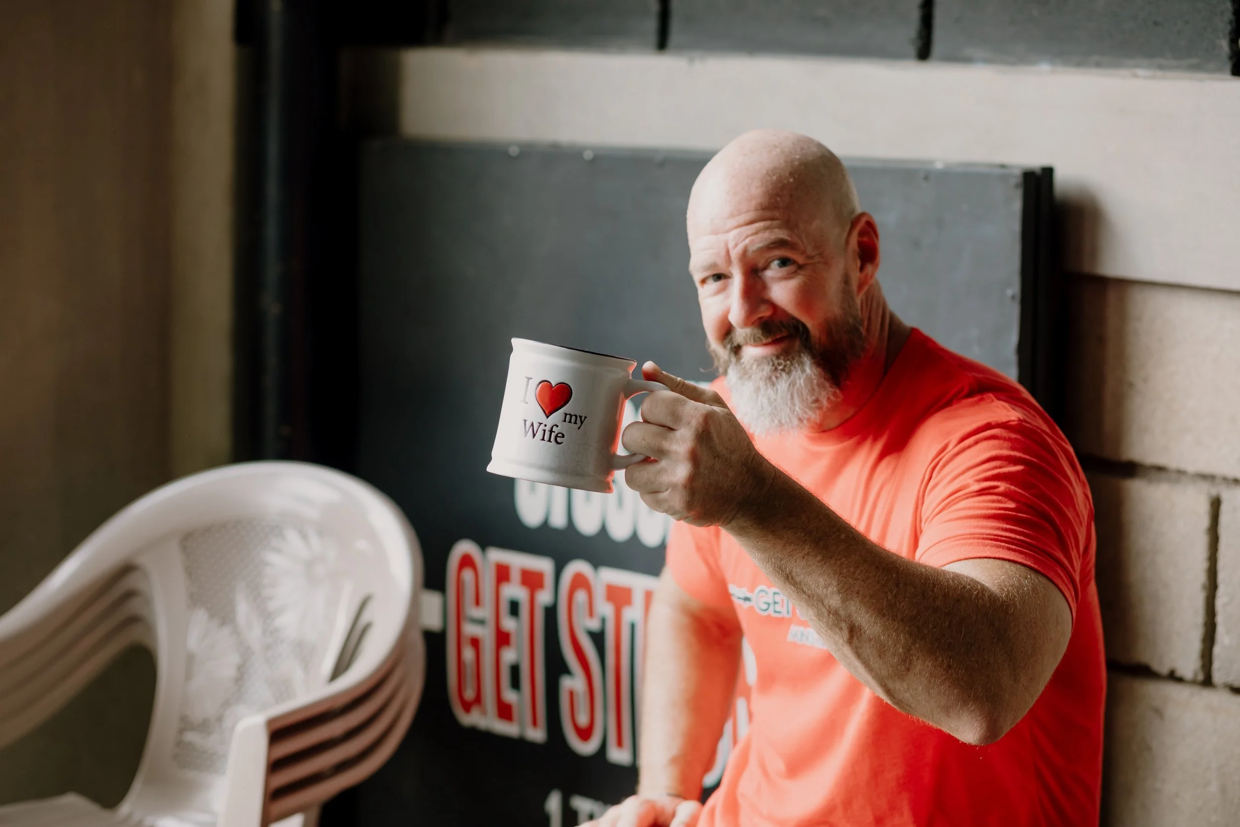 A man with a bald head and a beard, smiling and holding a white mug with a red heart and the words 'I love my wife' printed on it, sitting near a white plastic chair in a casual indoor setting.