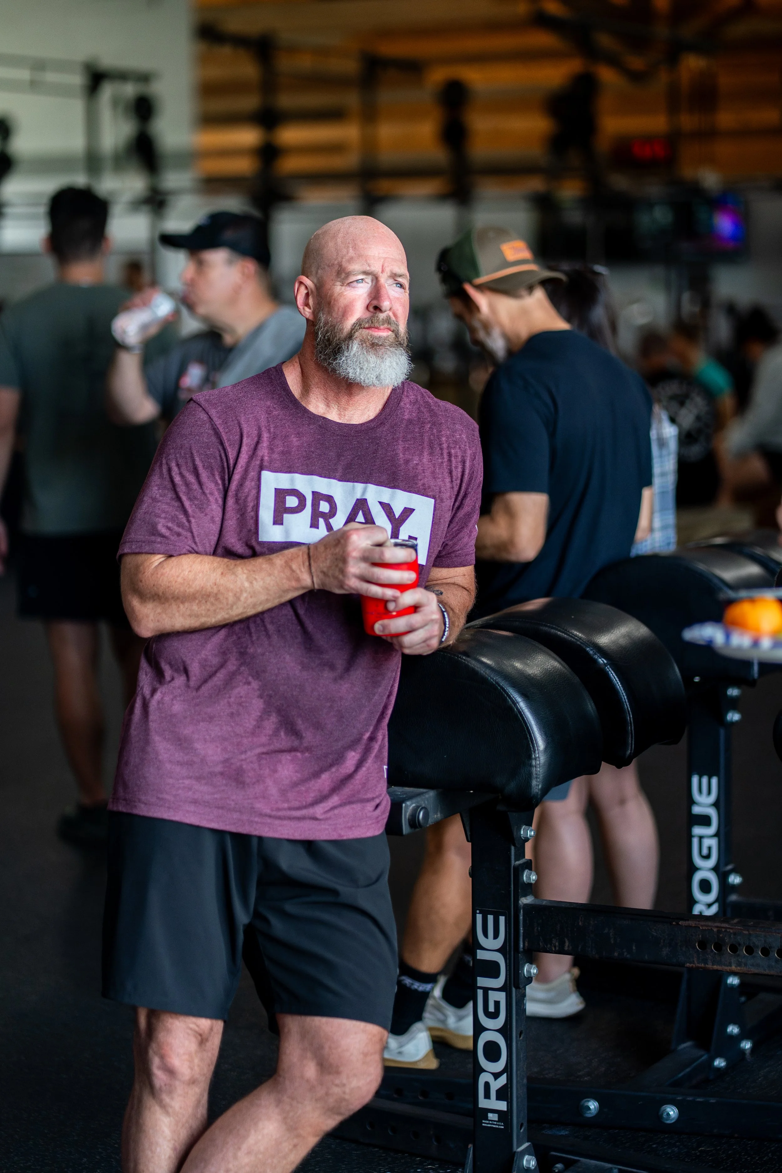 A man with a beard and bald head wearing a purple t-shirt with the word 'PRAY' on it, standing in a gym beside a workout bench, holding a red cup, with other people in the background. Some are drinking water, and the gym has wooden walls and fitness 