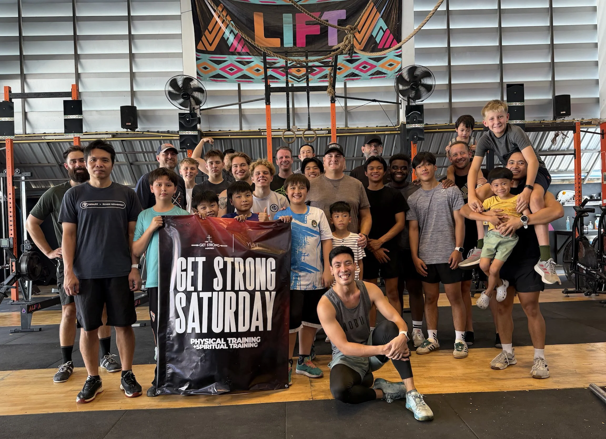 A group of children and adults in a gym or fitness center, holding a banner that reads 'Get Strong Saturday' with additional text about physical and spiritual training. They are smiling and standing in front of gym equipment and a colorful wall banner that reads 'LIFT.'