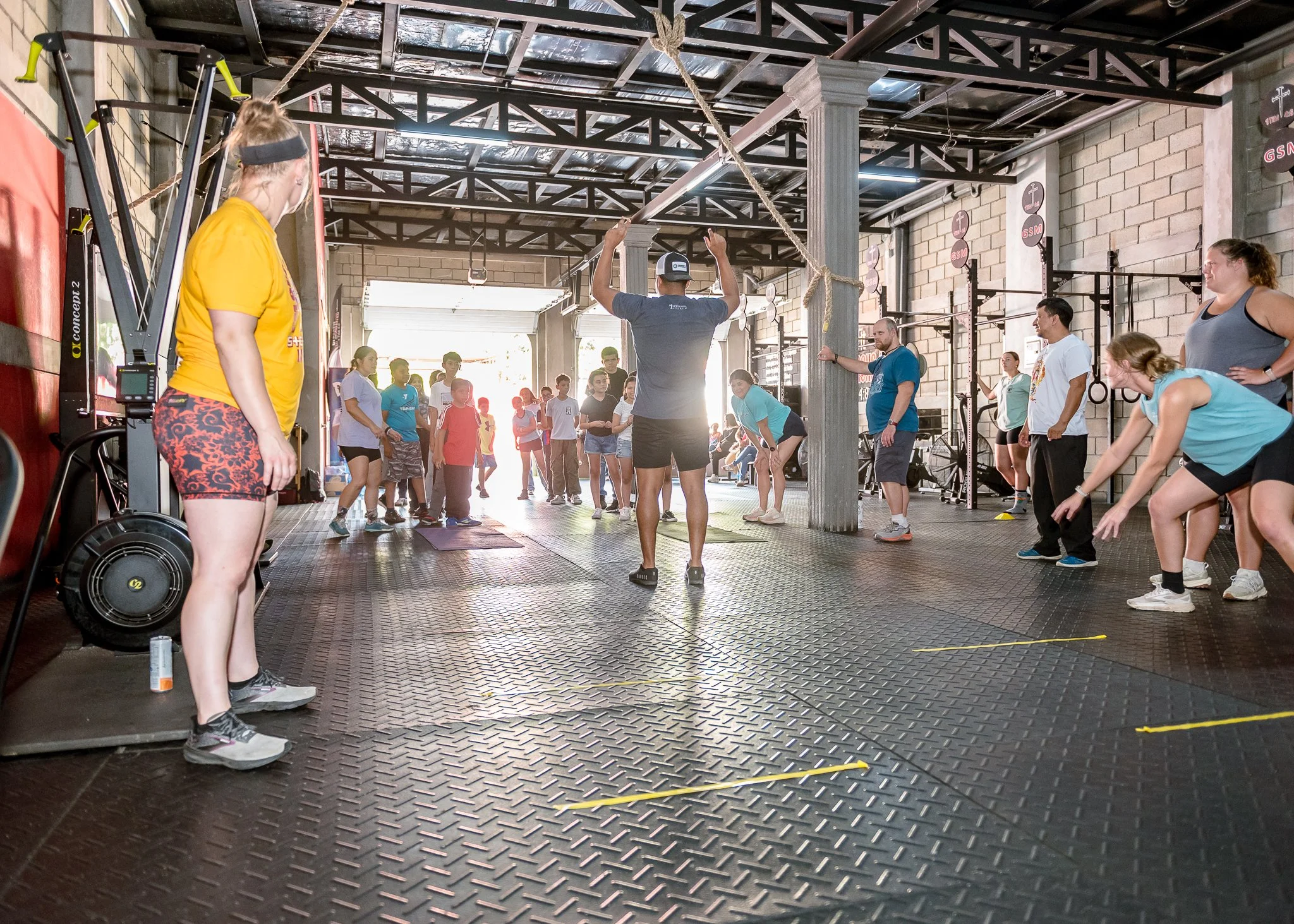 People participating in a fitness class at a gym, with an instructor demonstrating an exercise using a rope, and others following along, inside an industrial-style gym with brick walls and workout equipment.