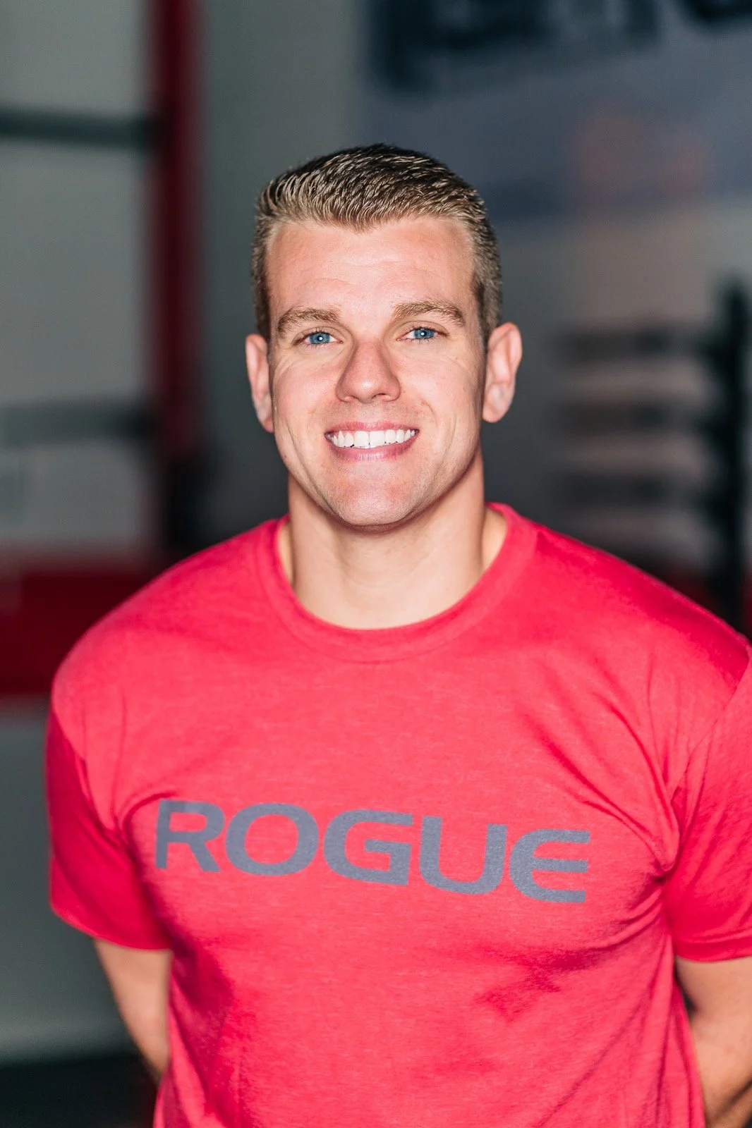 A smiling man with short hair in a red shirt with the word 'ROGUE' printed on it, standing in a gym.