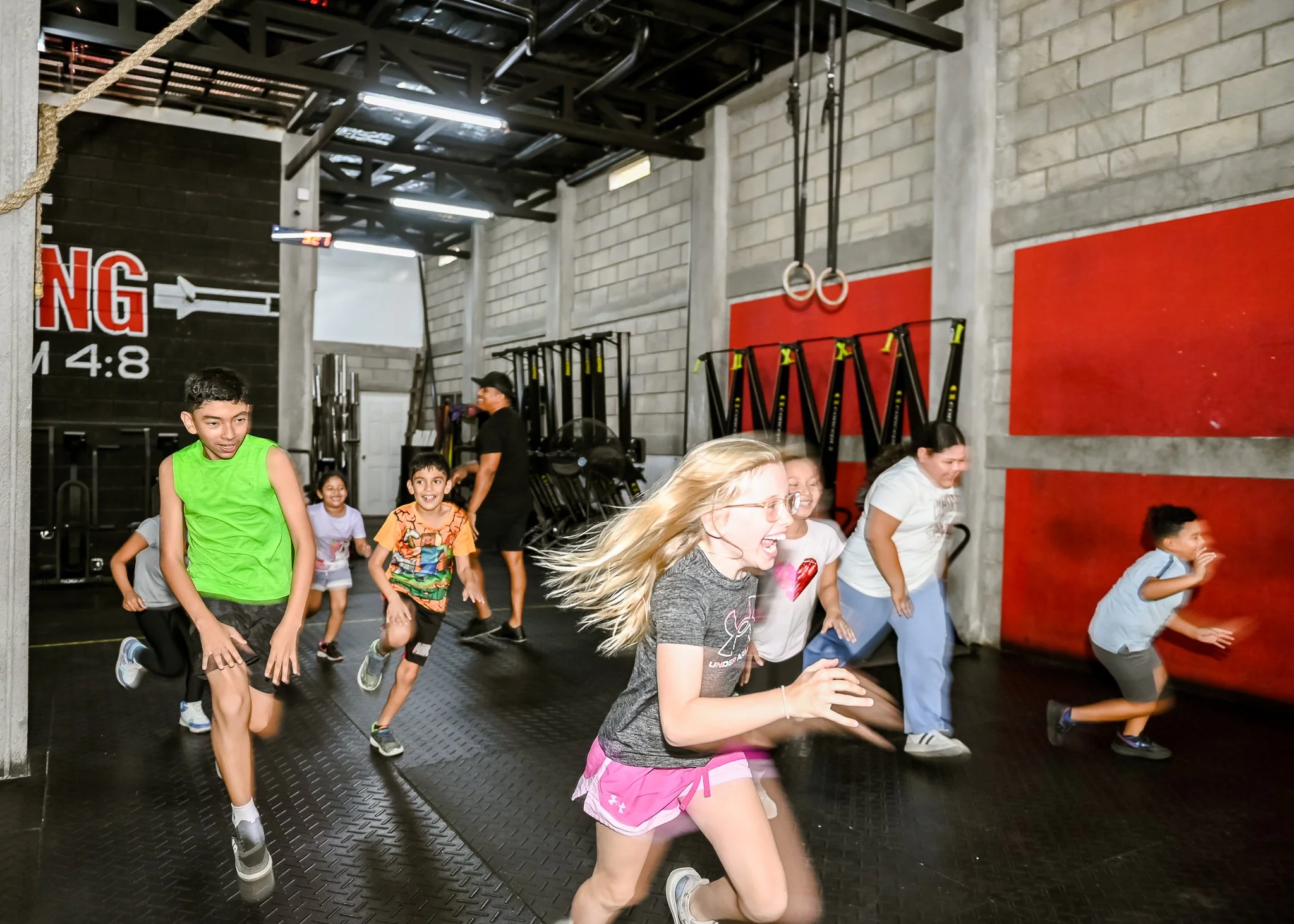A group of kids and women running through a gym with workout equipment in the background.