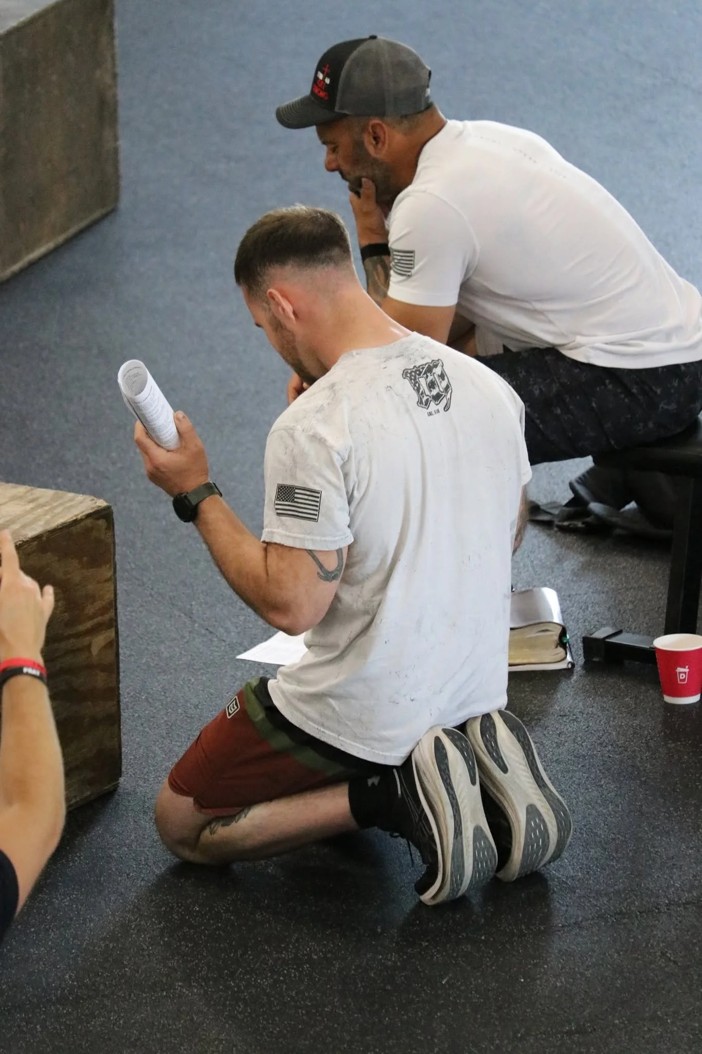Two men kneeling on a black speckled gym floor, praying. The man in the foreground wears a white T-shirt with American flags on the sleeve, maroon shorts, and sneakers. The man in the background wears a white T-shirt, black pants, and a black cap. Va