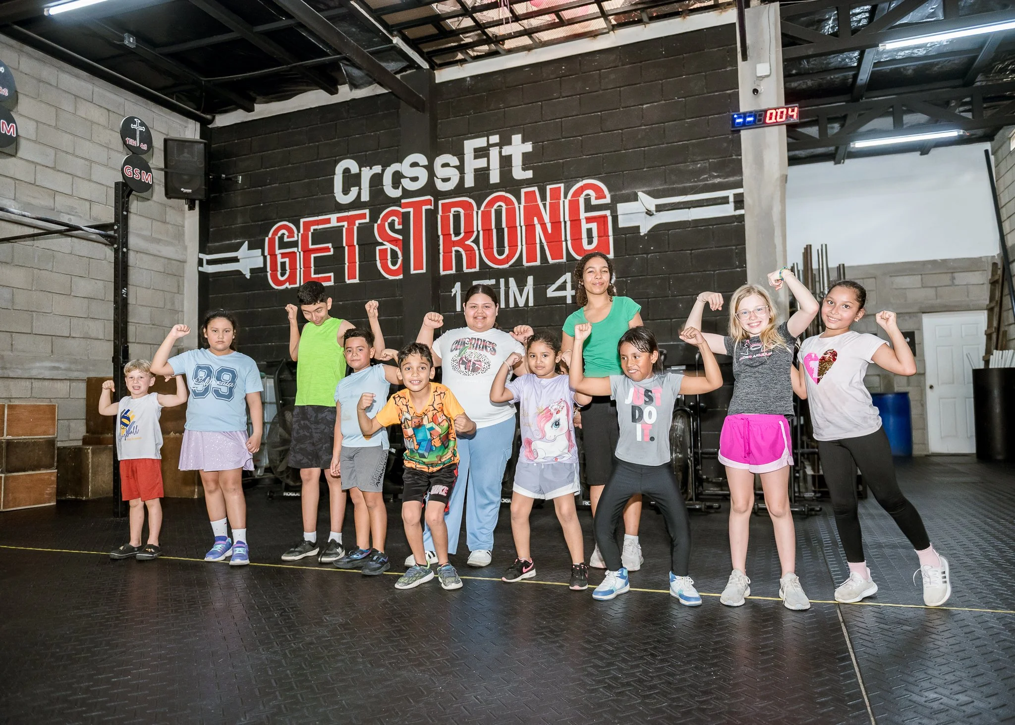Group of children and teenagers posing with their arms flexed inside a CrossFit gym. The wall behind them has the phrase 'CrossFit GET STRONG' painted on it, and a digital timer is visible in the upper right corner.