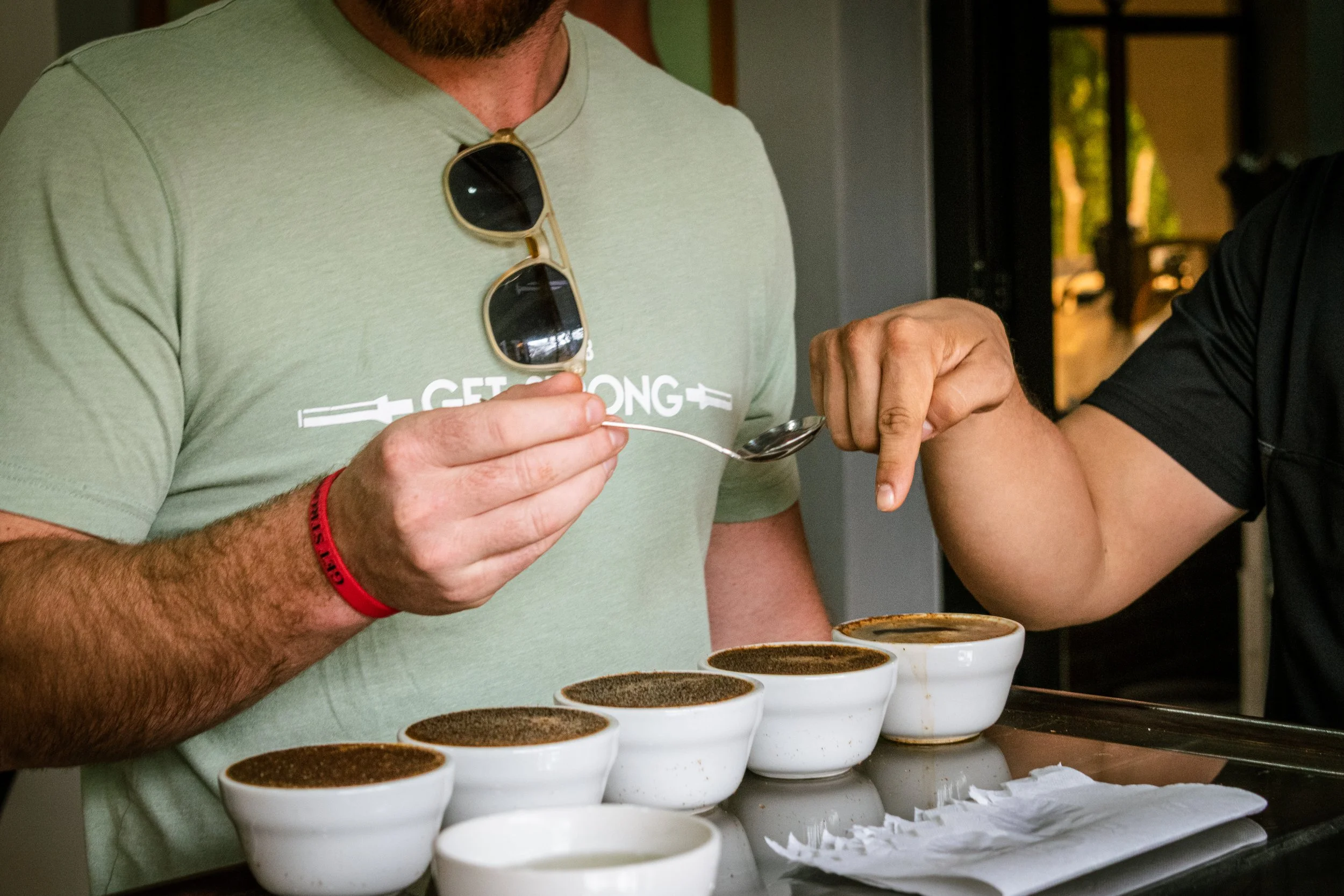 A person wearing a green shirt with sunglasses hanging from the collar, holding a spoon, and arm in arm with another person in black shirt, standing behind several cups of coffee or hot chocolate on a counter.