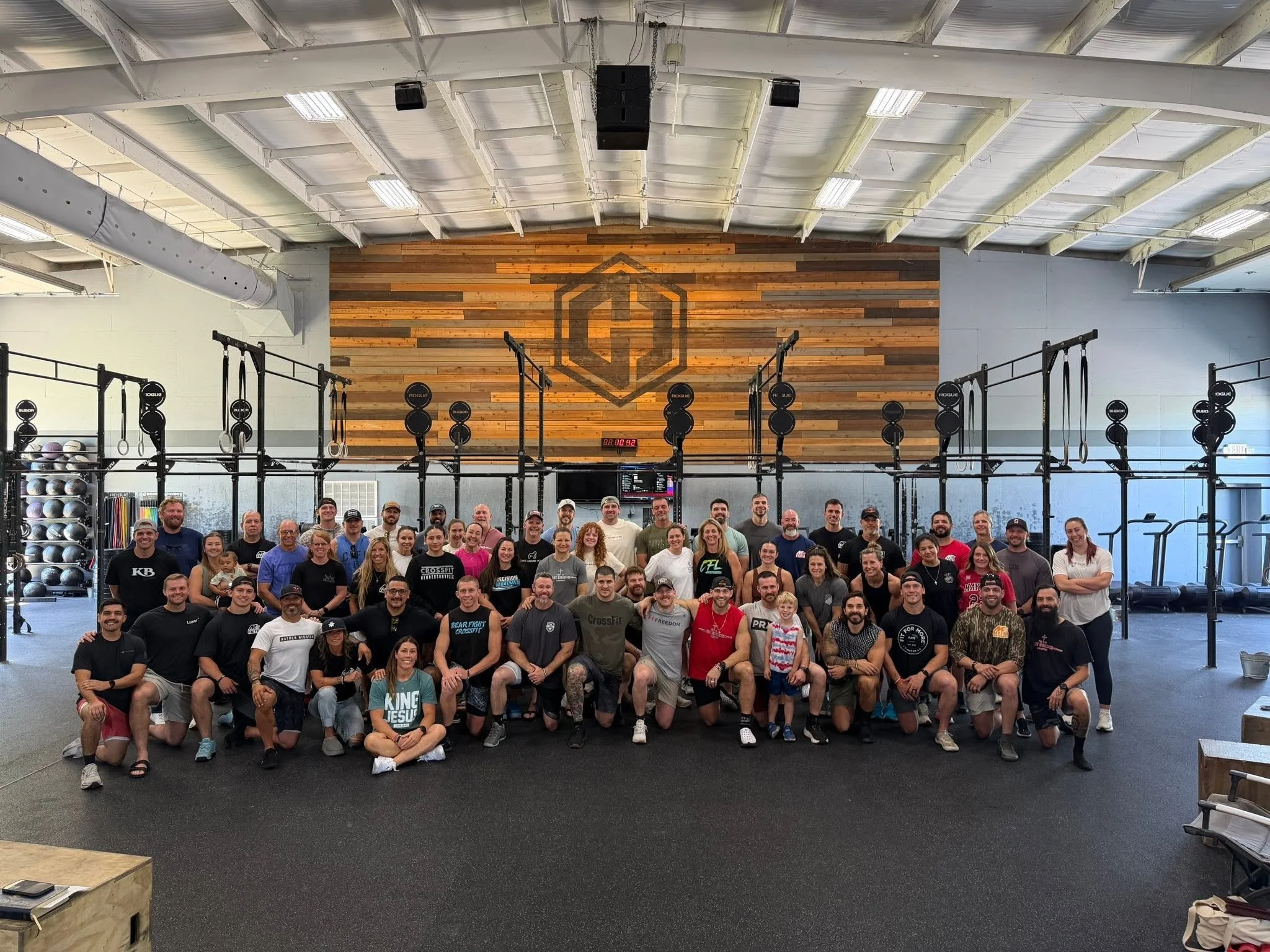 A large group of people, men and women, children, gathered inside a gym or fitness center for a group photo. The background features fitness equipment and a wooden wall with a large logo. The group is smiling and appears to be celebrating or marking 