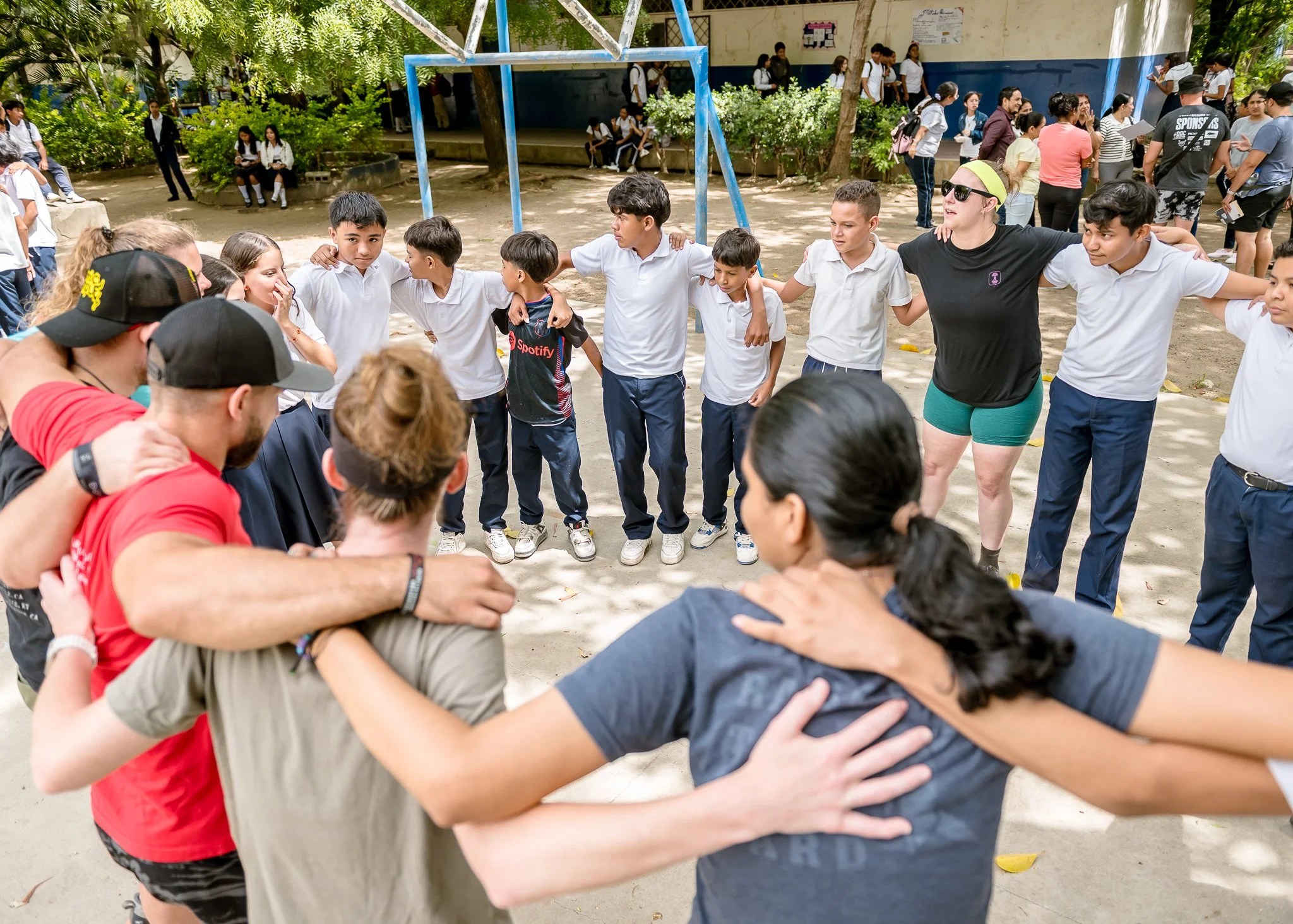 A group of students and adults gather outdoors in a circle, with their arms around each other's shoulders. In the background, more students are sitting on benches or walking. The setting appears to be a schoolyard with trees and a playground structure, on a sunny day.