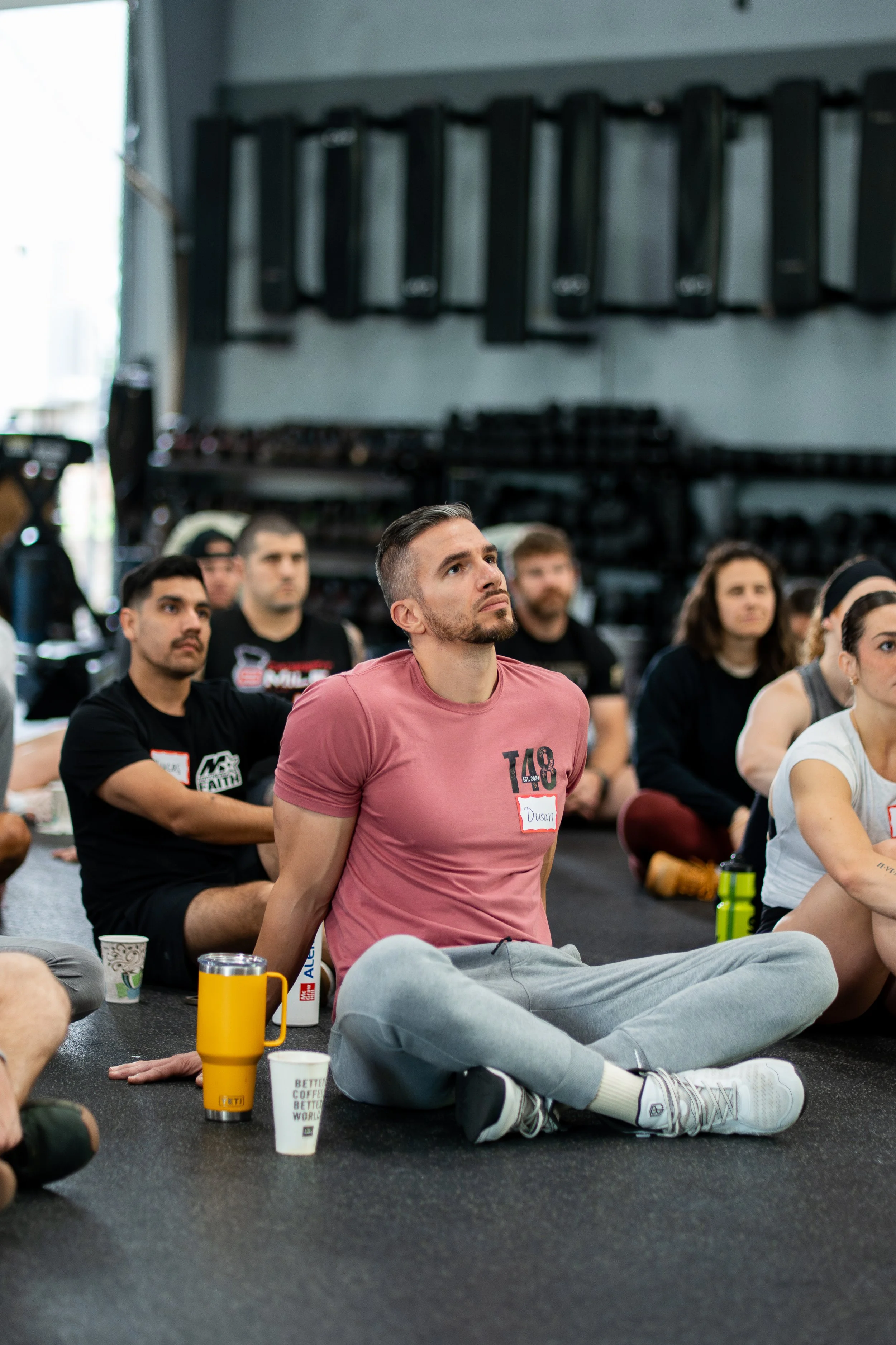 People sitting on the floor in a fitness or yoga class, some with name tags, surrounded by water bottles and coffee cups.