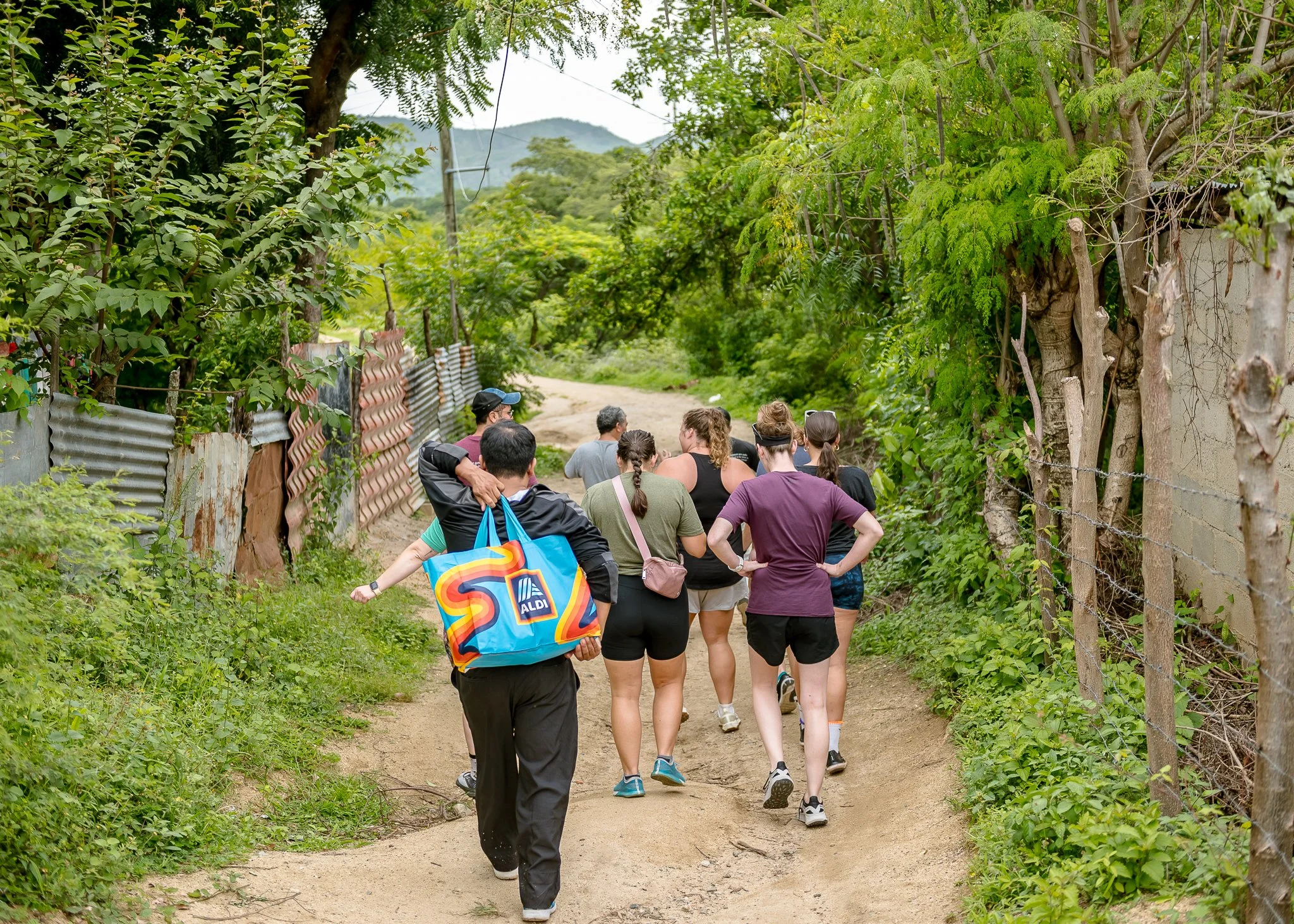 A group of people walking along a dirt path surrounded by green trees and foliage, with mountains in the background.