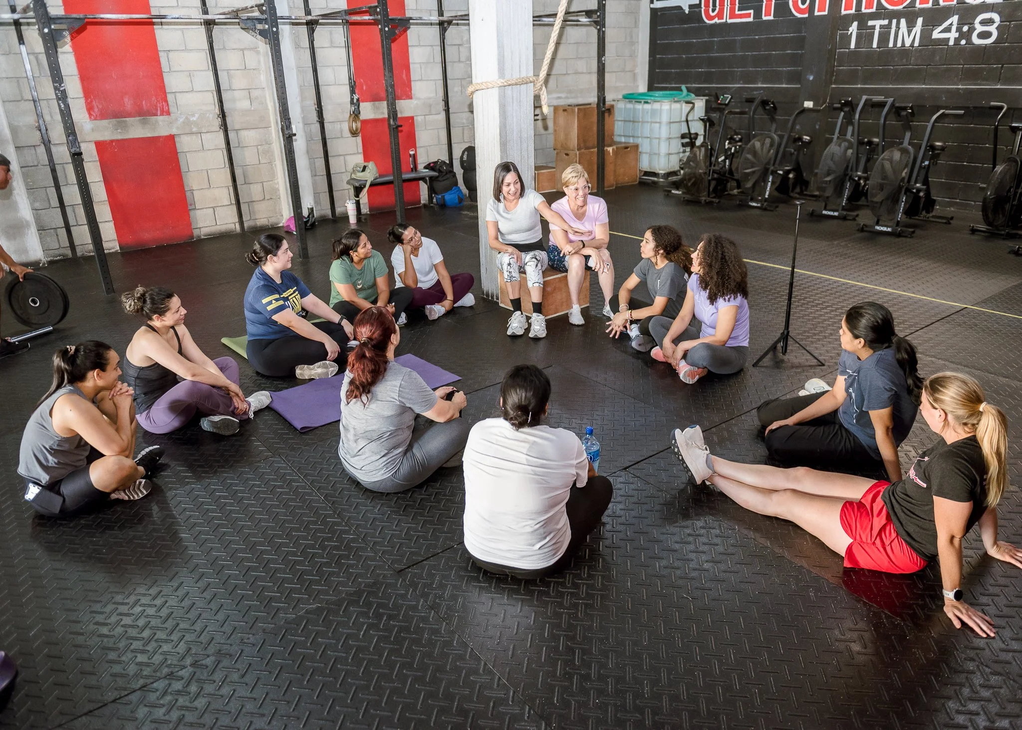 A group of women at a CrossFit gym participating in a fitness class. Some women are sitting on the floor, while two women are sitting on a wooden box at the center, smiling and talking. The gym has fitness equipment, black rubber flooring, a black wa