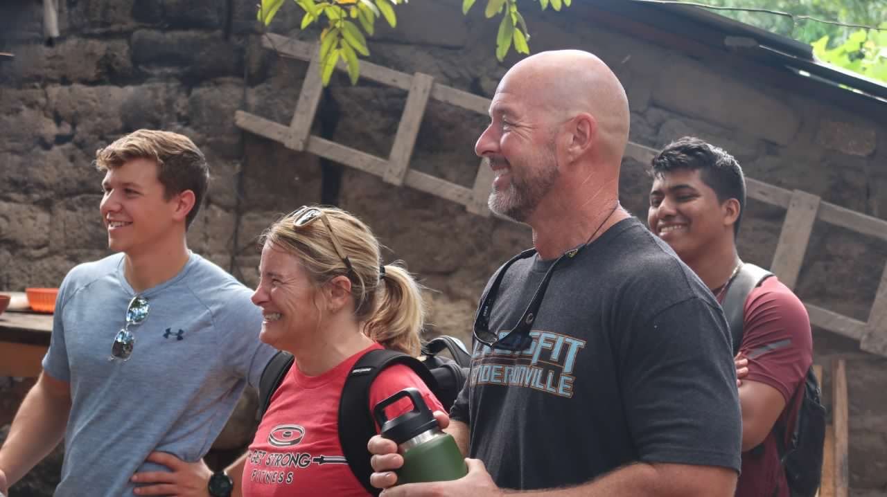 A group of five people smiling and enjoying a moment outdoors. They are standing next to a stone wall with wooden beams and greenery above. The group includes both men and women, some wearing backpacks and casual athletic clothing.