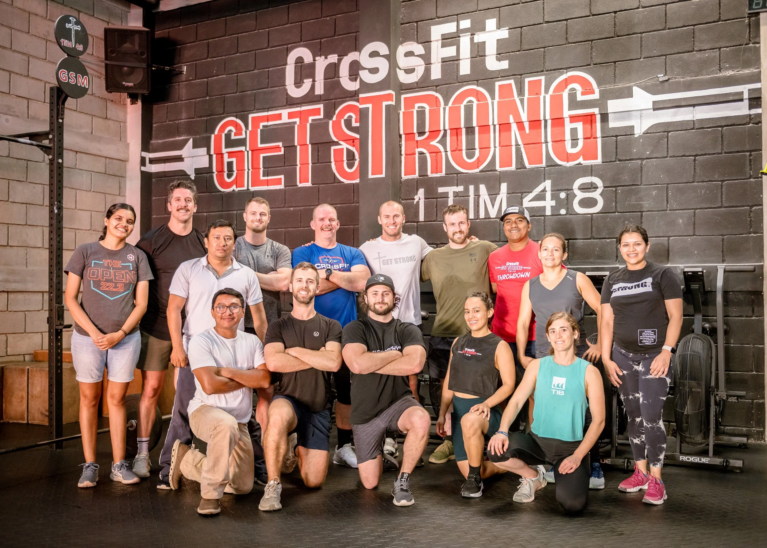 Group of people posing inside a CrossFit gym with a large sign on the wall reading "CrossFit GET STRONG" and a timer showing 4:48.