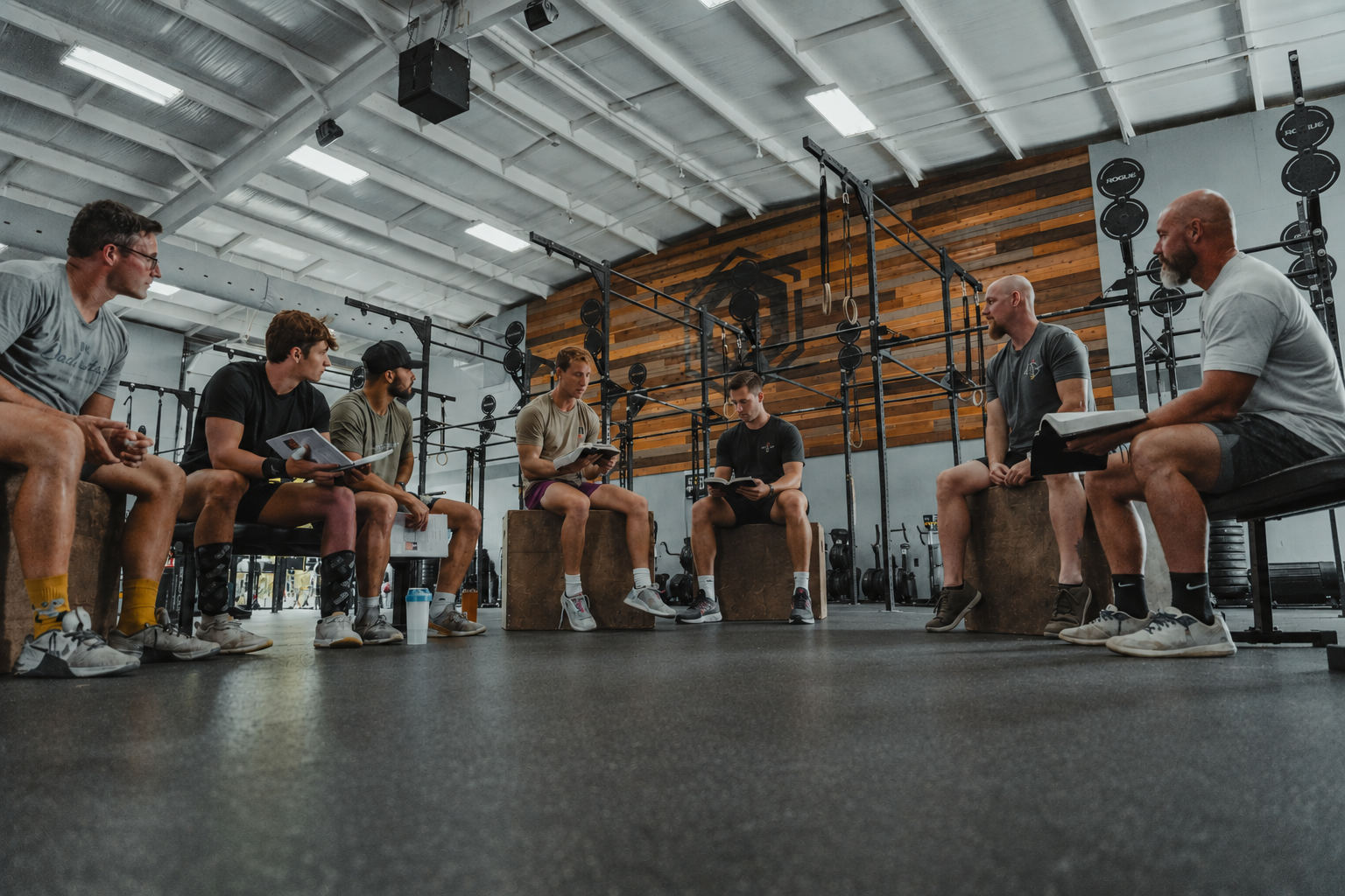 Group of seven men sitting on workout benches and plyometric boxes in a gym, participating in a fitness class or training session, reading from books or guides, with gym equipment and a wooden wall in the background.