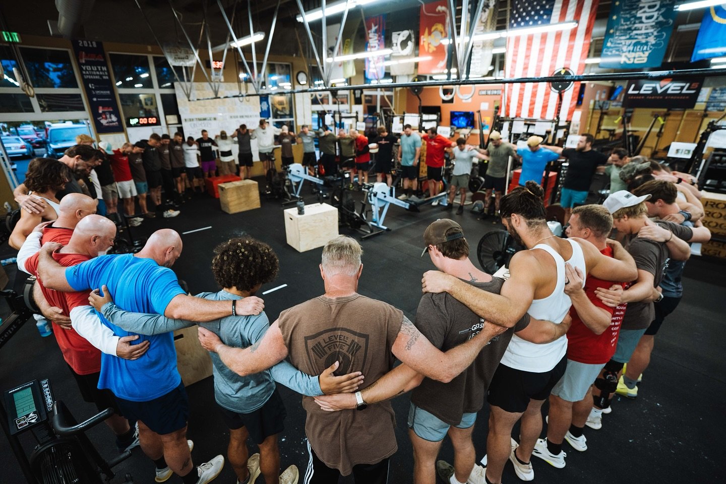 Group of people participating in a team-building exercise at a gym, standing in a circle with their arms around each other's shoulders.