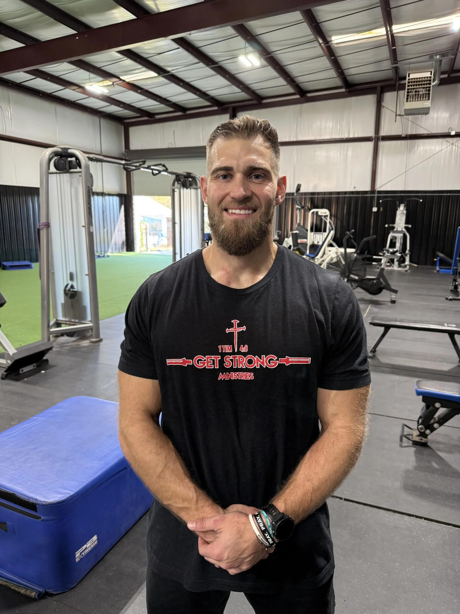 Smiling man with a beard wearing a black T-shirt that says 'Get Strong Ministries' in a gym with exercise equipment and a blue padding mat.