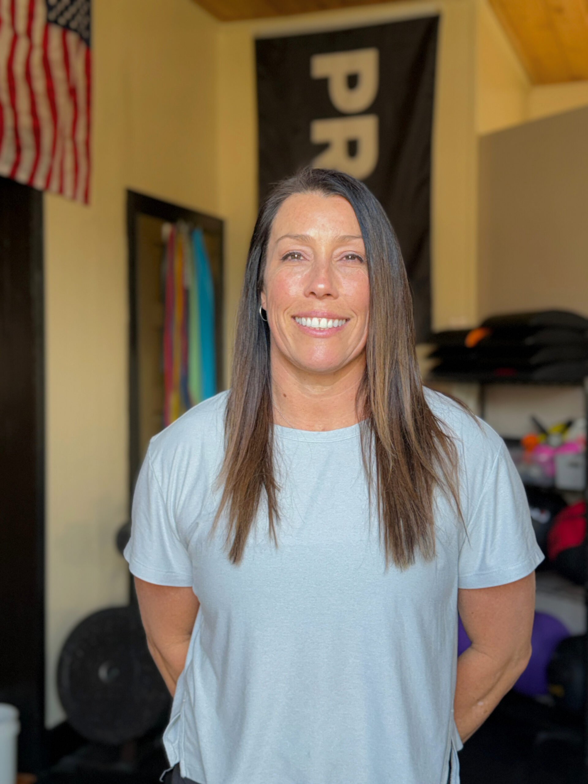 A woman with long brown hair, smiling, wearing a light gray t-shirt, standing in a room with workout equipment, barbell, and resistance bands in the background.