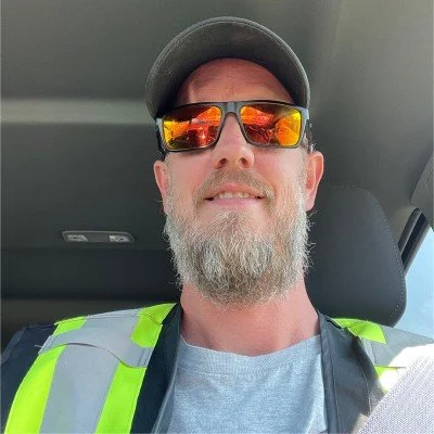 A man with a gray beard wearing sunglasses, a baseball cap, and a safety vest sitting inside a vehicle.