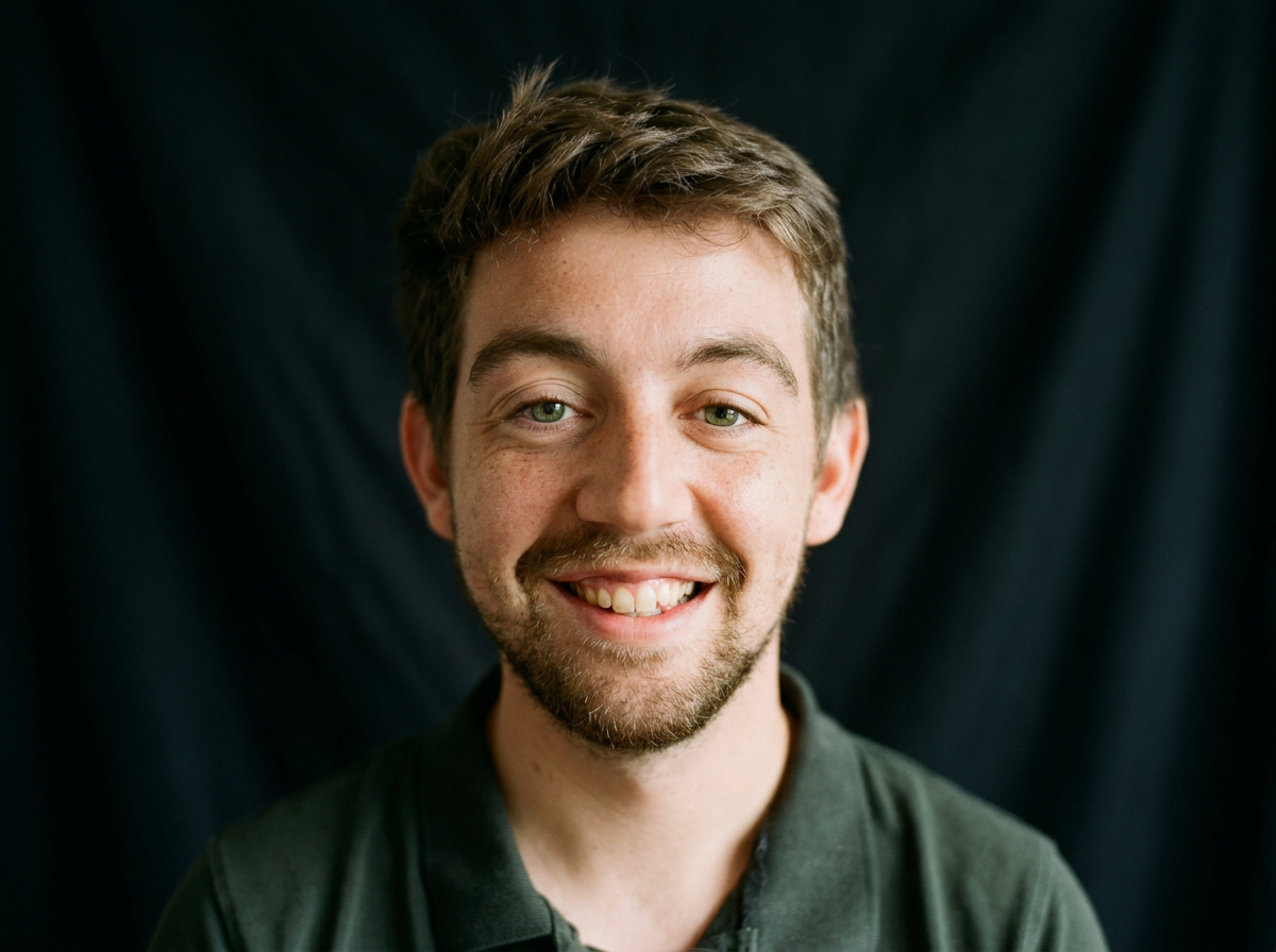 Close-up of a young man with brown hair, green eyes, and a beard, smiling and showing teeth, against a dark background.