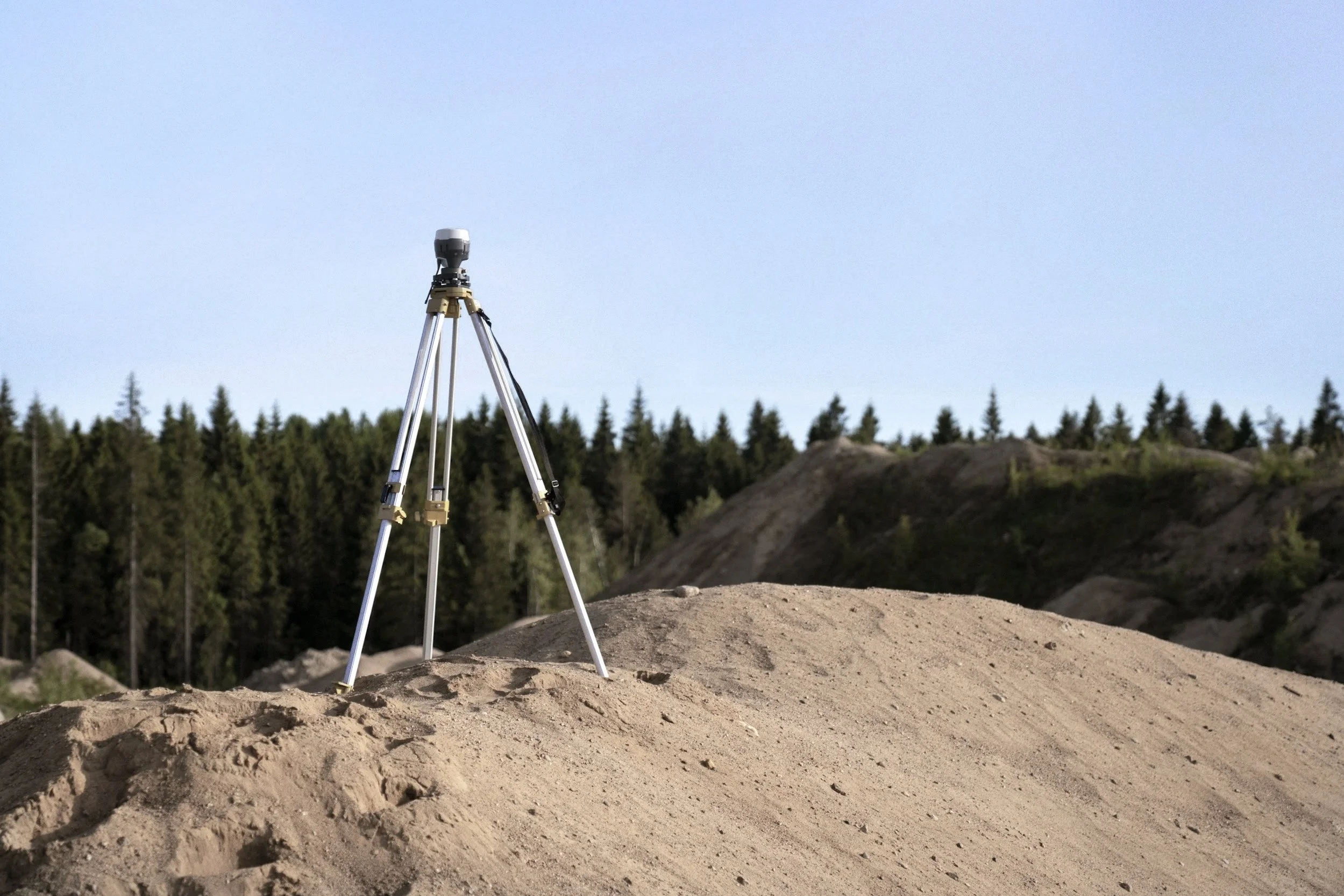 A surveying tripod with a mounted total station on a dirt mound in a rural outdoor area with a forest and blue sky in the background.