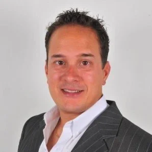 Portrait of a man with short dark hair, wearing a dark striped suit and white shirt, smiling against a plain light background.