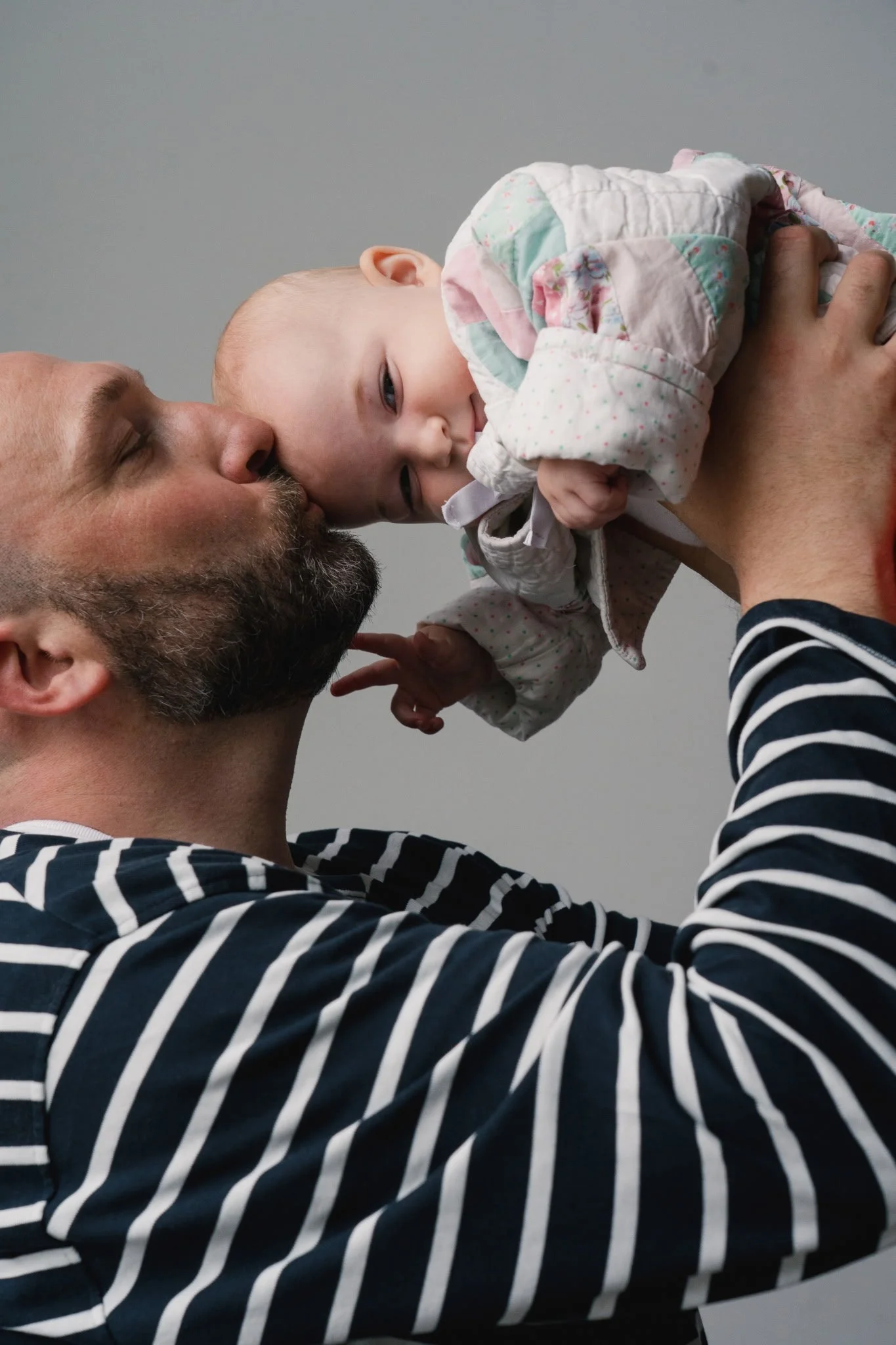A man with a beard and striped shirt holding a baby girl with a floral quilted jacket, the man kisses the baby's forehead on a plain gray background.