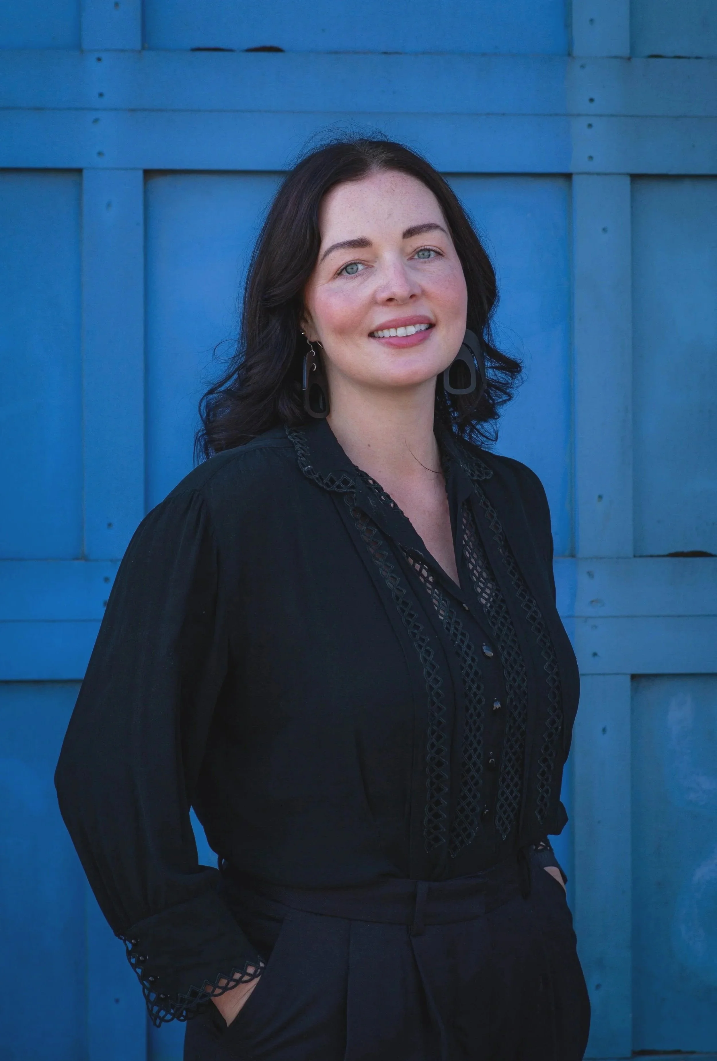 A woman with black hair and blue eyes smiling at the camera, wearing a black blouse with lace detailing, standing in front of a blue wooden wall.