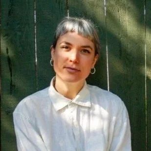 A woman with short gray hair and hoop earrings, wearing a white collared shirt, standing in front of a green wooden fence with sunlight and shadows.