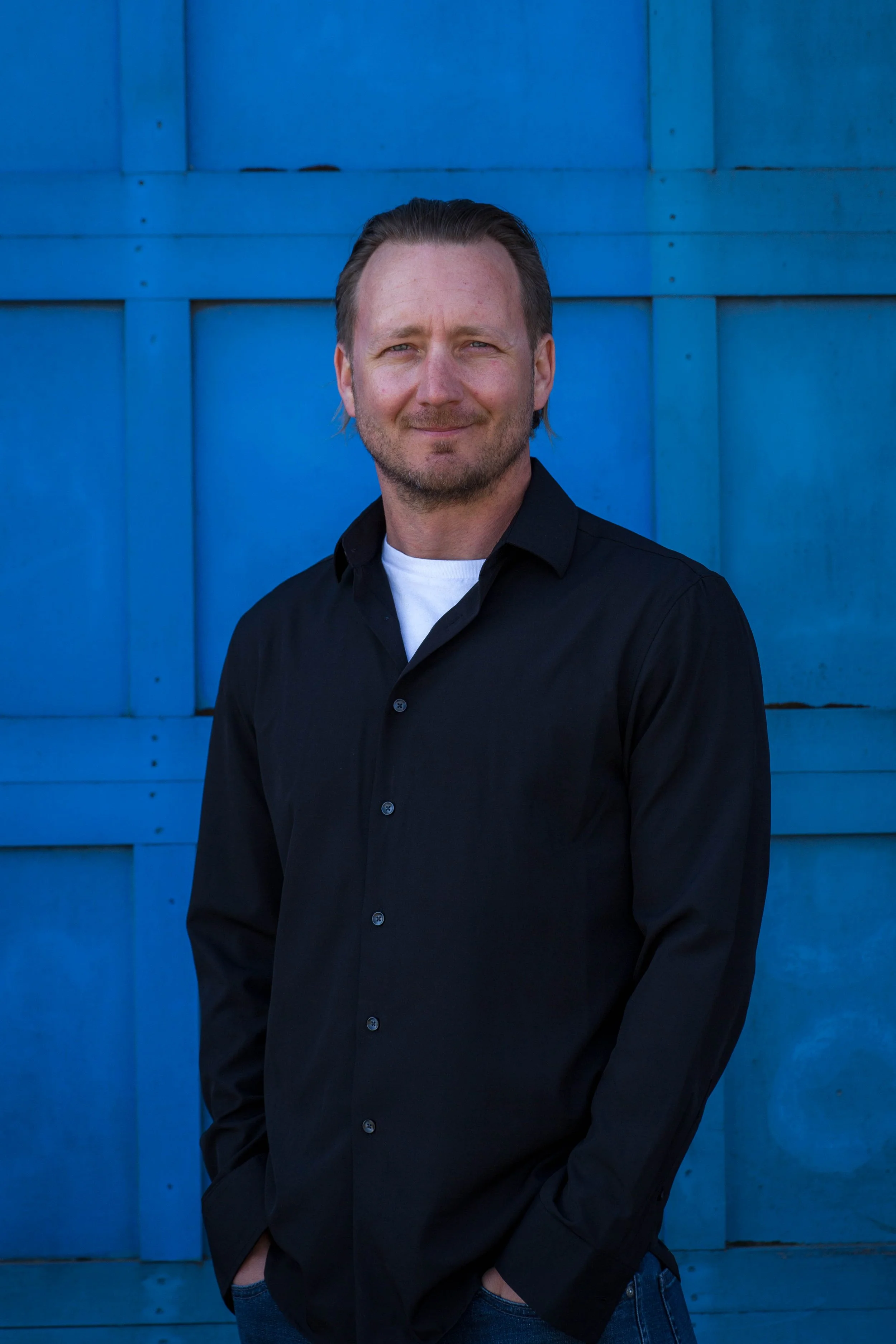 A man standing in front of a bright blue wooden wall, wearing a black shirt and jeans.