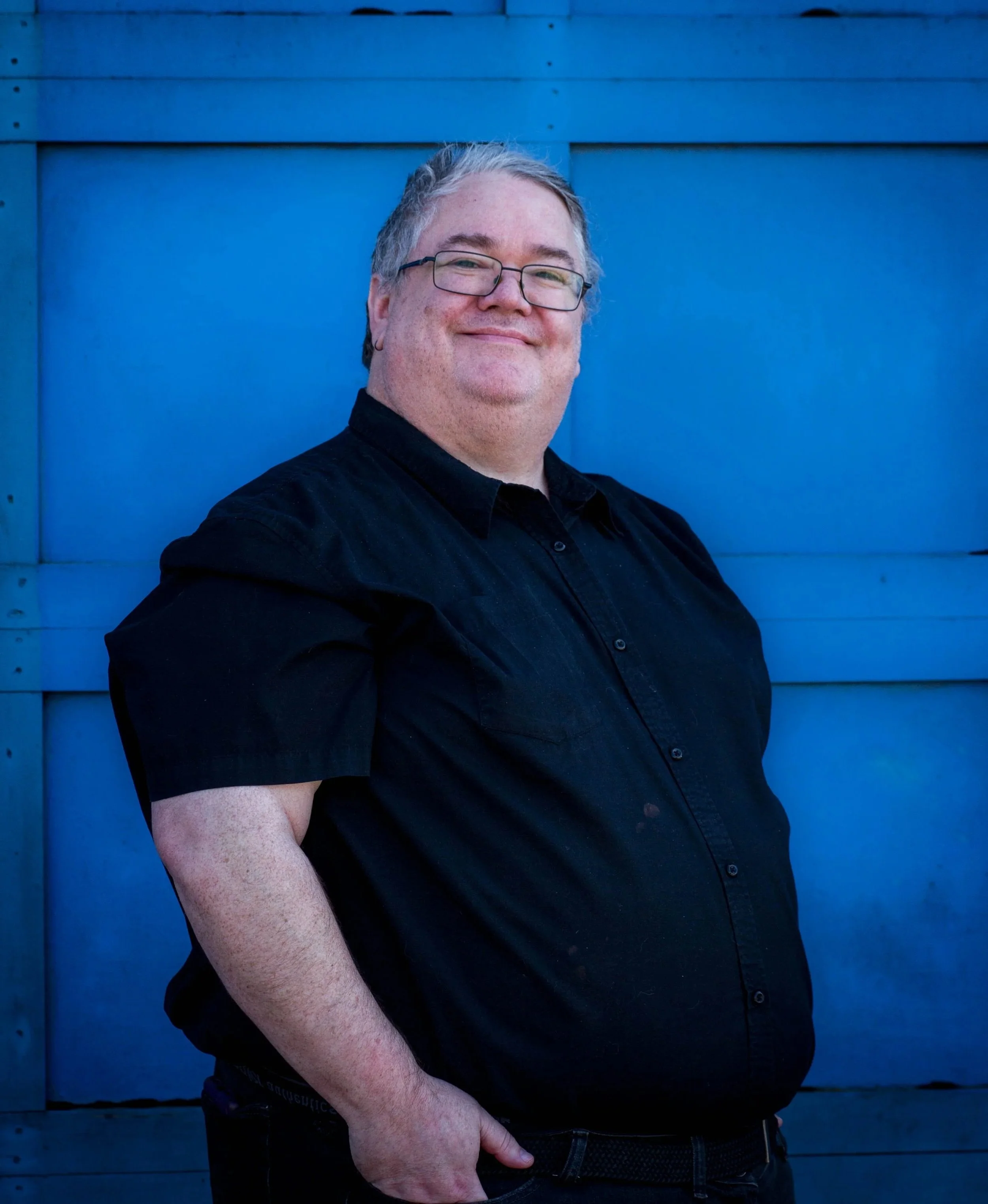 A man with glasses and gray hair smiling, wearing a black shirt, standing in front of a blue wooden wall.