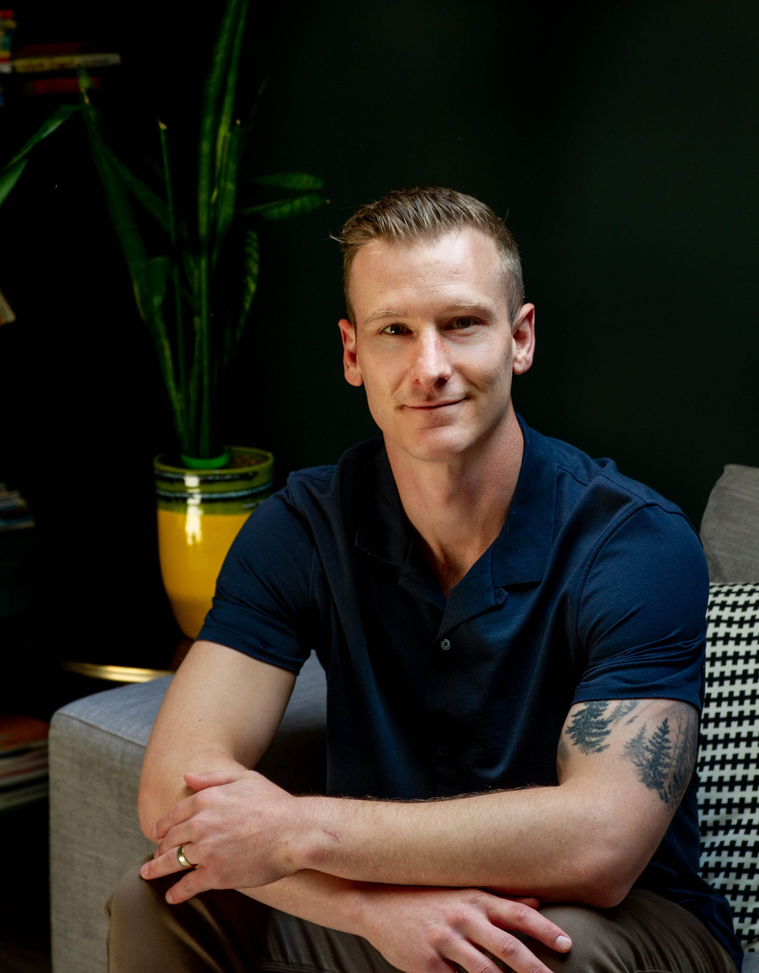 A man with short light brown hair, fair skin, and a tattoo of trees on his left arm, sitting indoors against a black wall, wearing a navy blue polo shirt, with a plant in a yellow pot and a bookshelf in the background.
