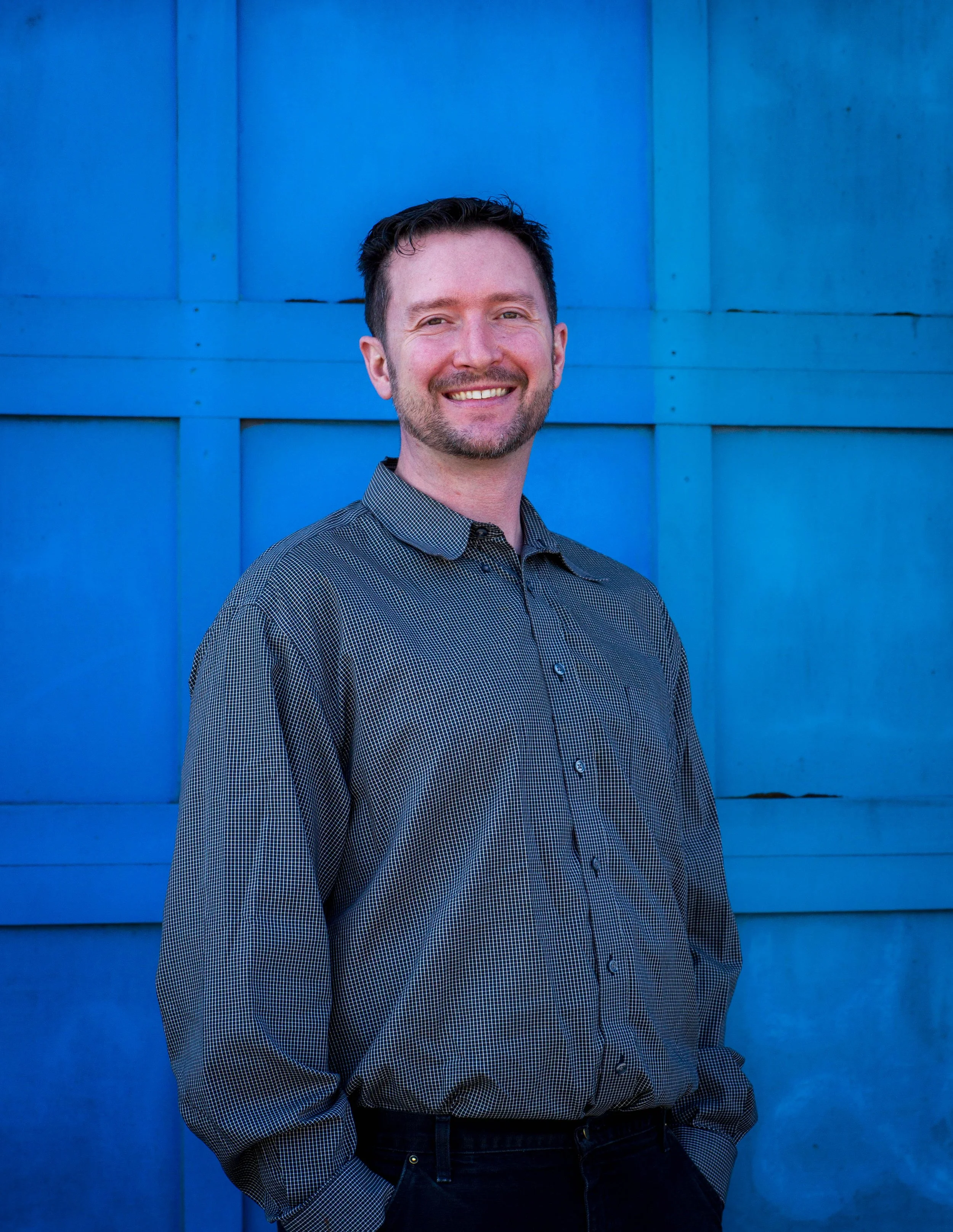 A smiling man with short dark hair and a beard, wearing a checkered shirt, stands against a blue wooden wall.